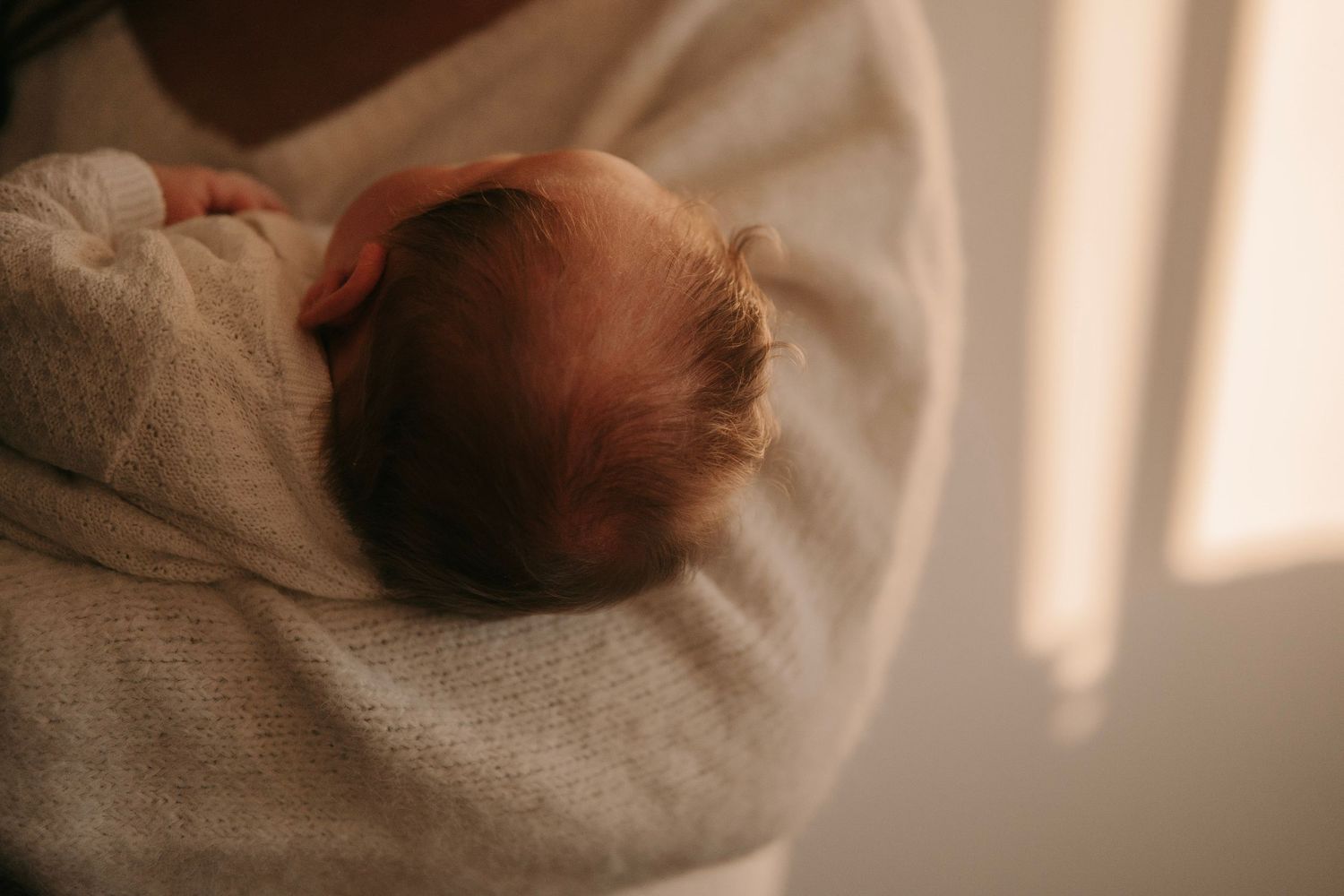 Close-up of a newborn baby with fine hair resting on a parent's shoulder wrapped in a white blanket.