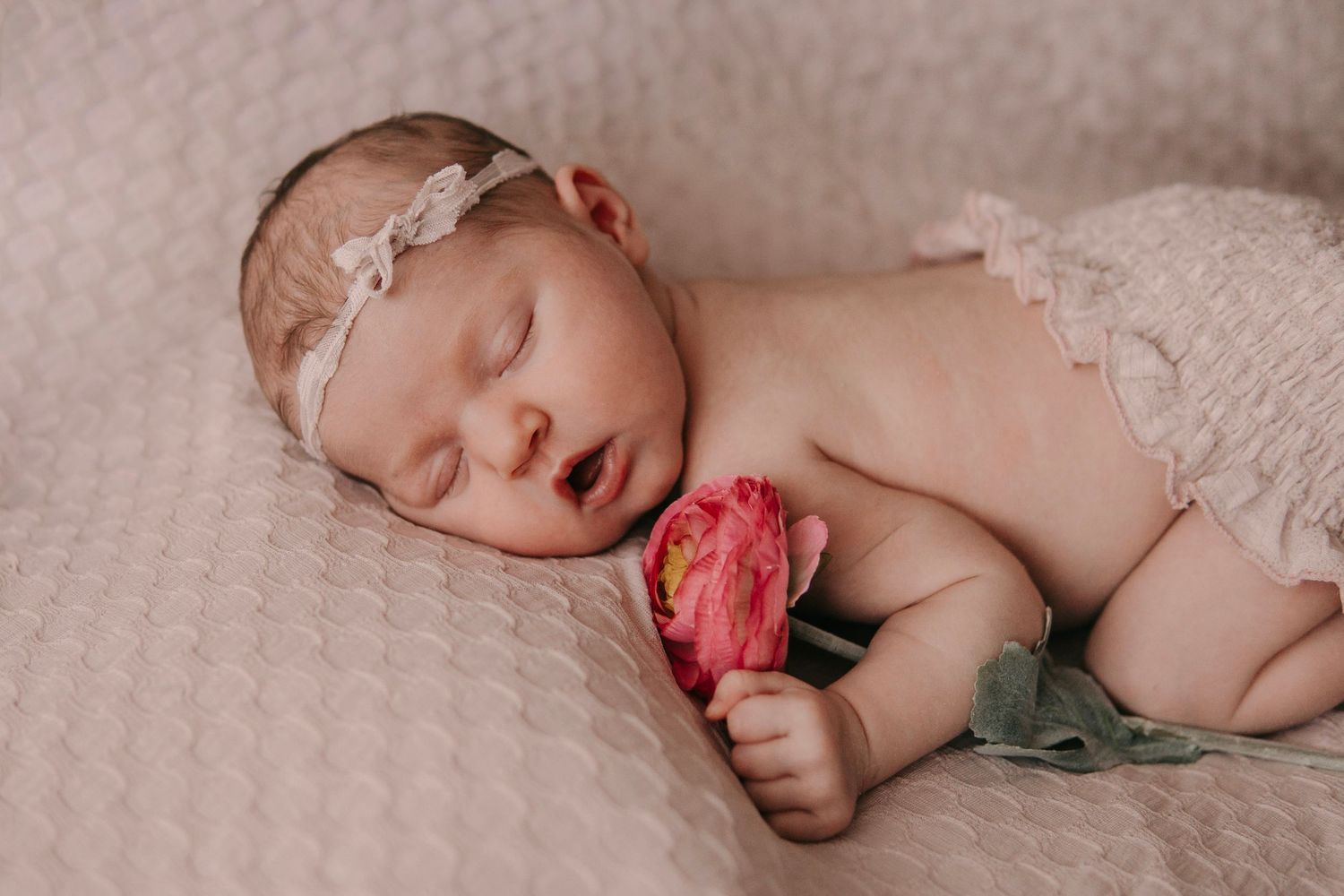 Sleeping newborn baby girl in pink lace outfit holding a pink flower, wearing a delicate floral headband.