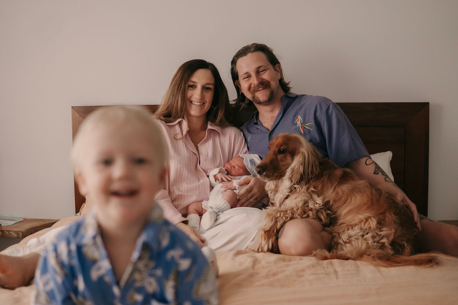 Happy family relaxing on a bed with a newborn baby, golden retriever, and toddler in a cozy home.