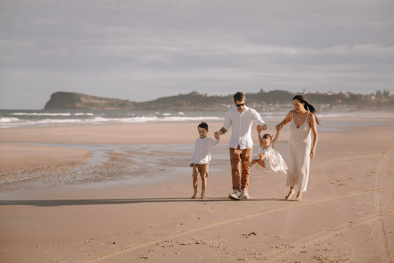 Family of four walking hand in hand on a sandy beach at sunset.