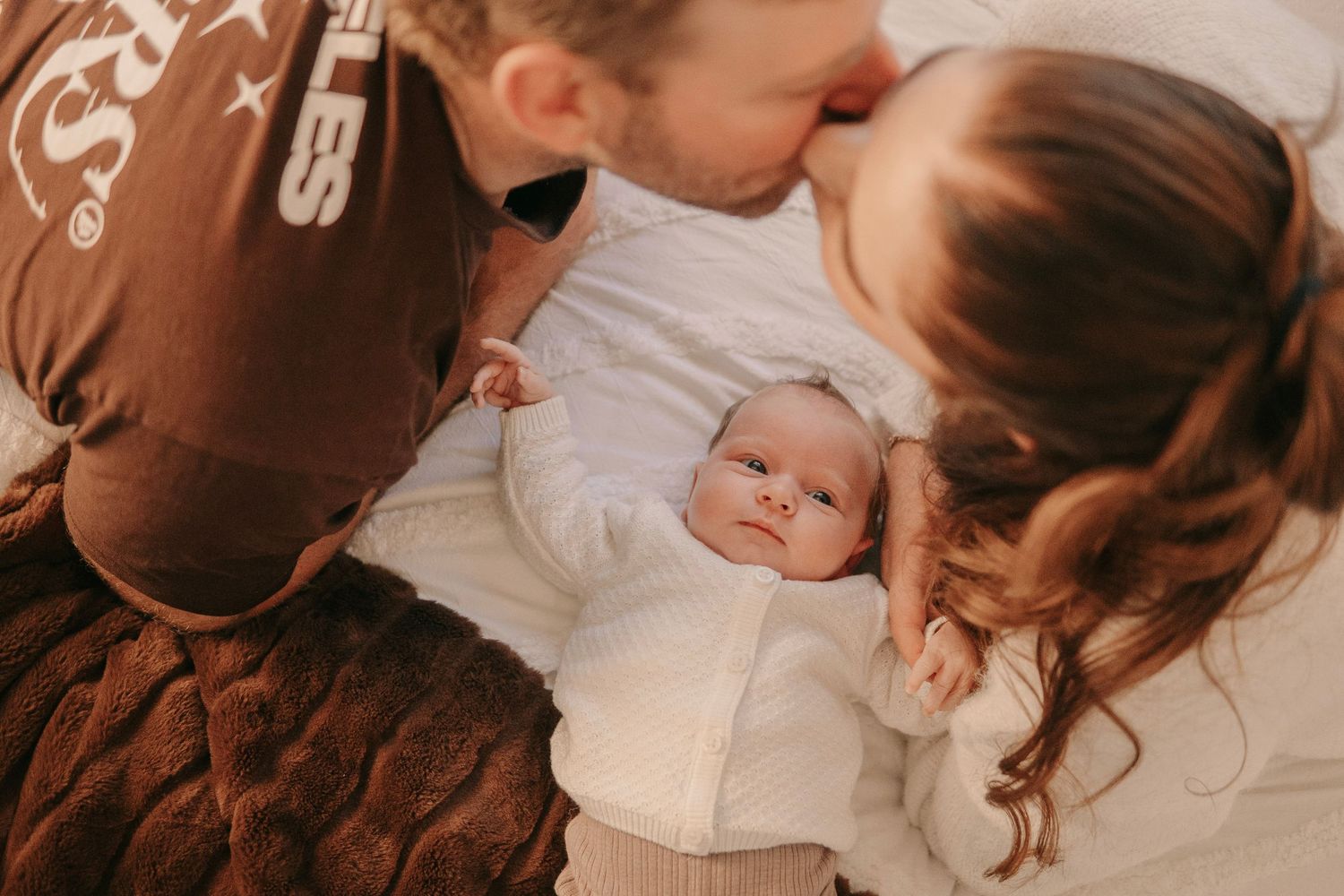 Parents kissing over their newborn baby wrapped in a white knit blanket.