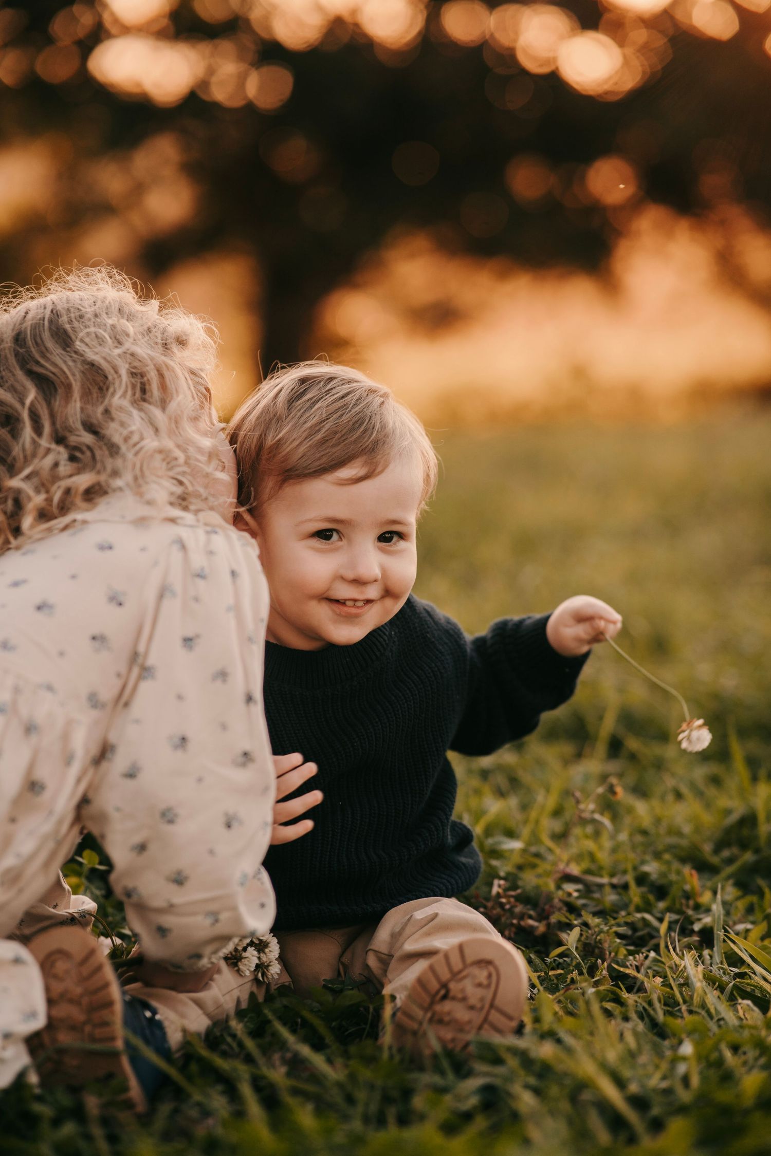 Cute toddler boy smiling at camera while sister hugs him in a golden sunset field.