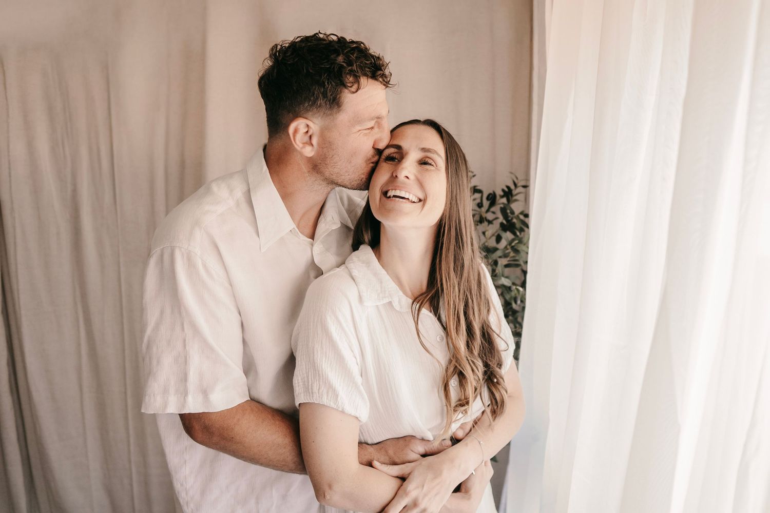 A couple in white outfits share a tender moment, man kissing woman on cheek while she smiles warmly.