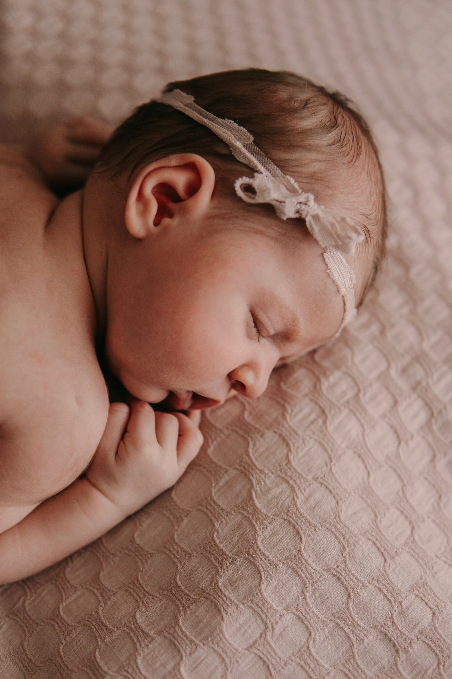 Sleeping newborn baby girl wearing a delicate white bow headband, resting peacefully on a soft cream textured blanket.