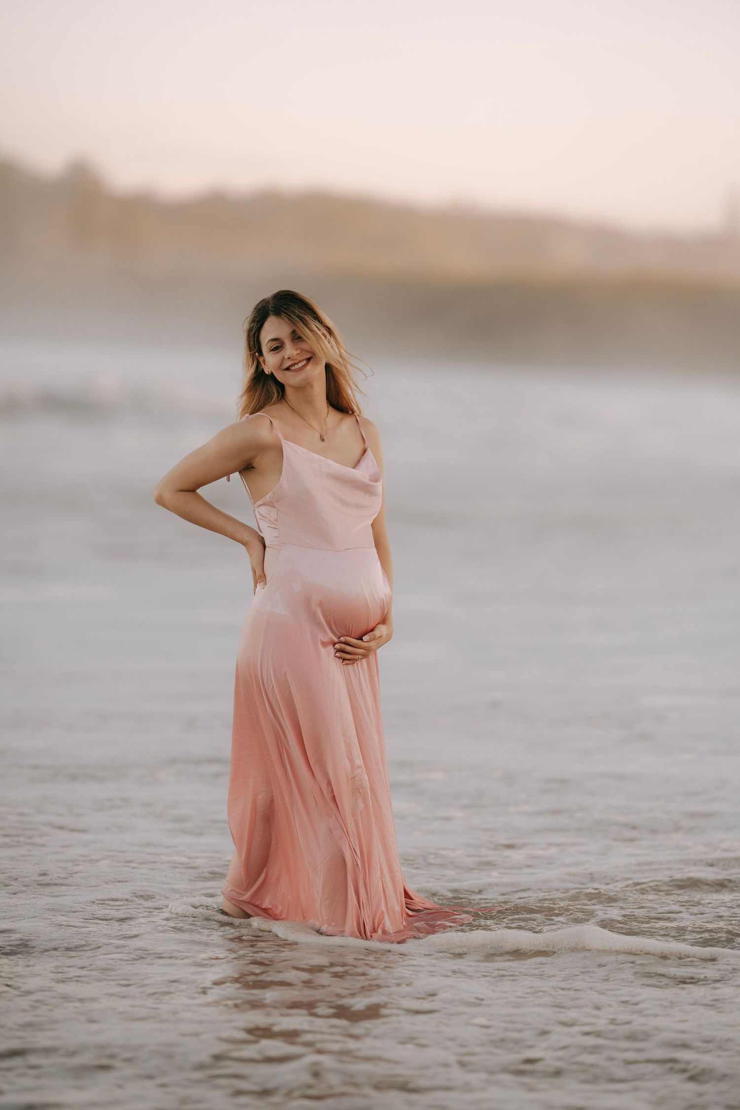 Smiling pregnant woman in pink dress standing in ocean waves at sunset.