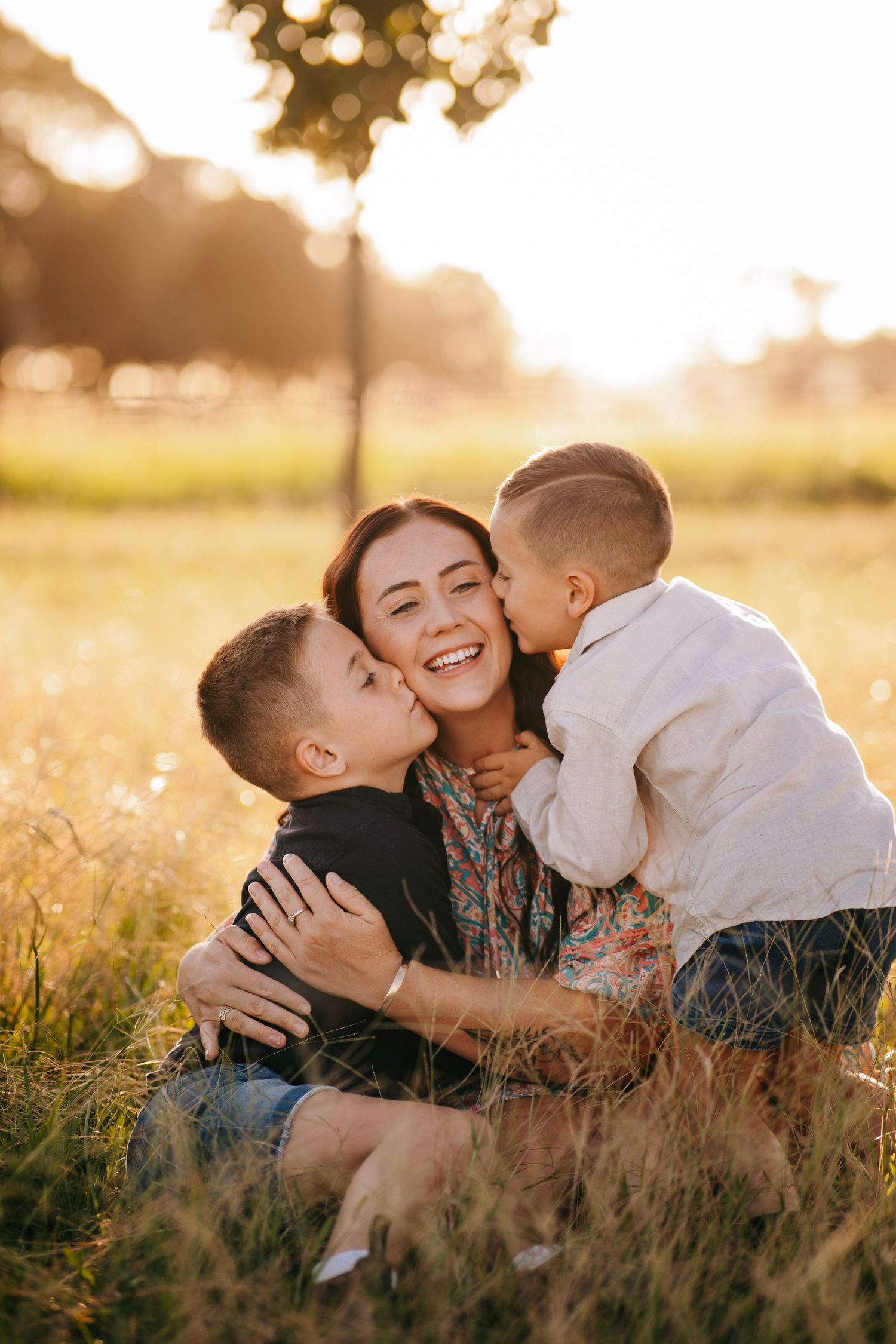 A smiling mother being kissed on both cheeks by her two young sons in a sunny field.