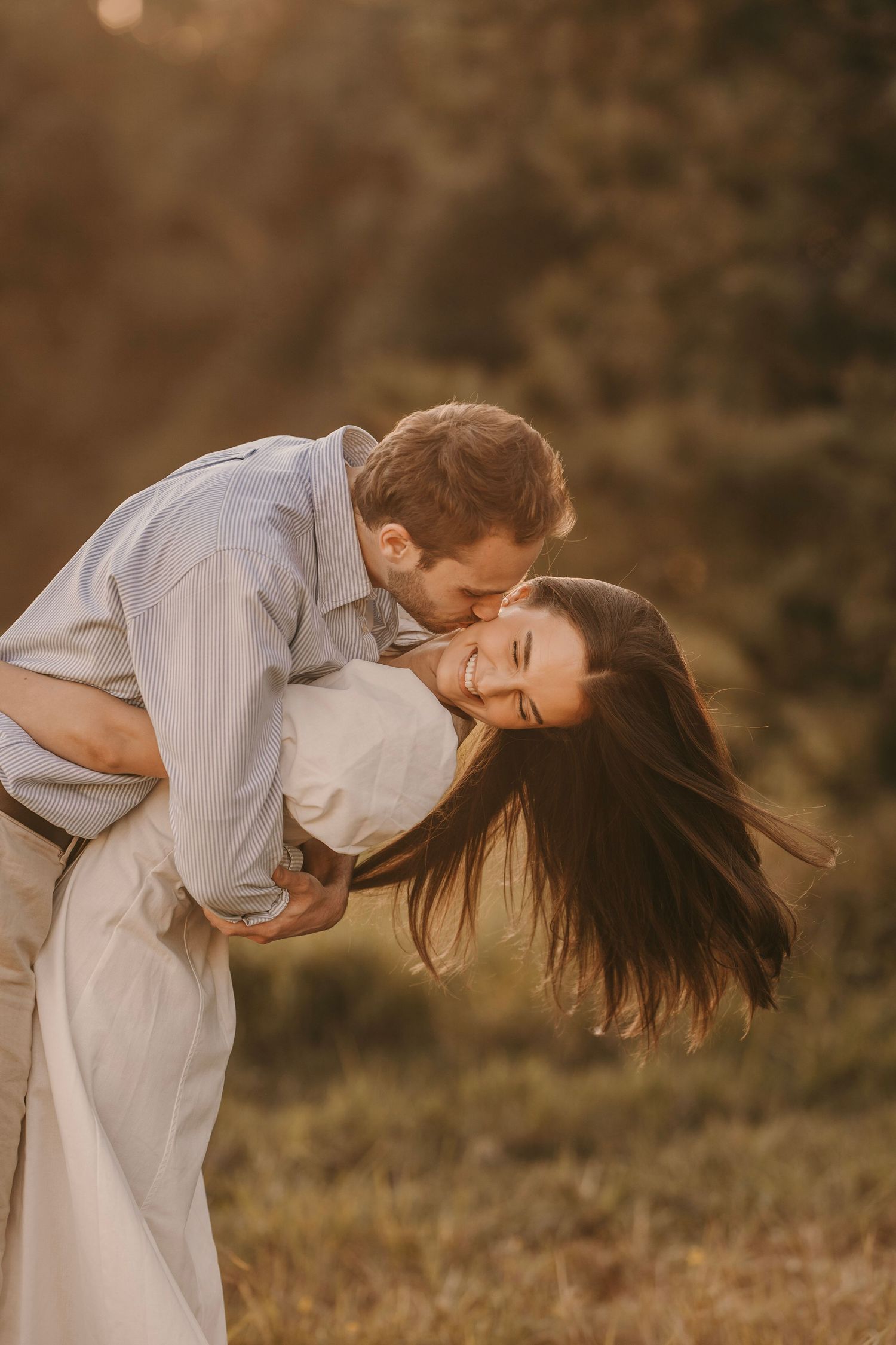 Man dipping and kissing laughing woman in a sunlit forest clearing during golden hour.