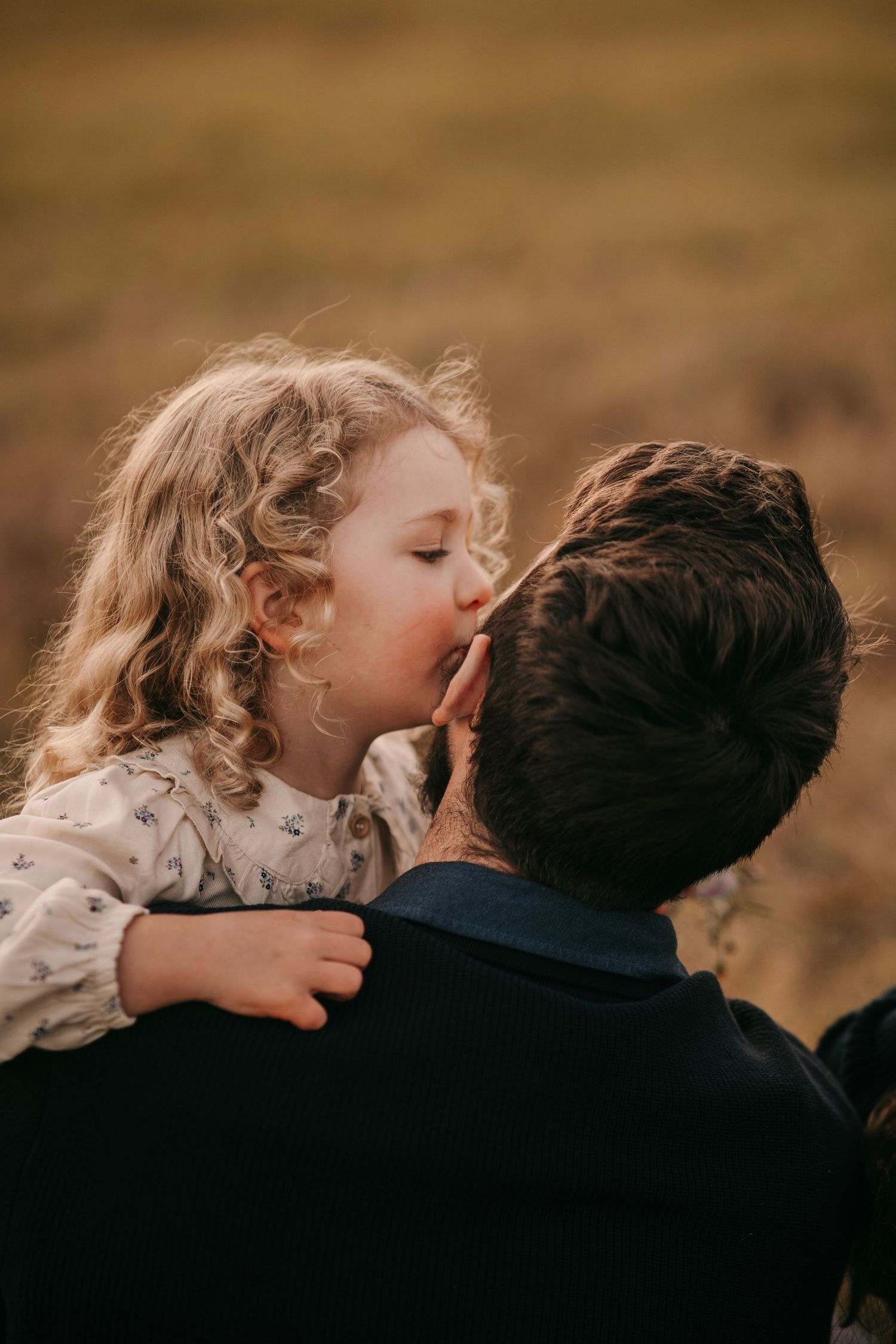 A young curly-haired girl gives her dark-haired father a sweet kiss on the cheek outdoors.