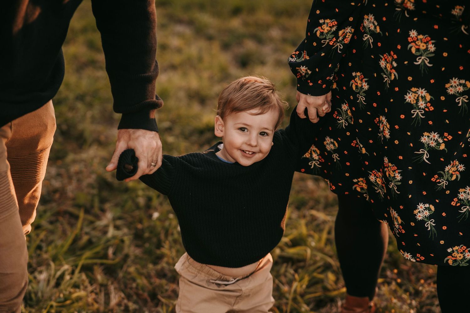 A smiling toddler boy holds hands with parents while walking on a grassy field at dusk.