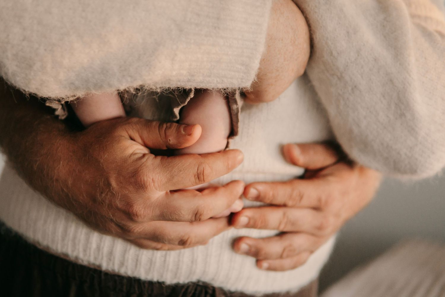 Close-up of fathers hands gently supporting newborn baby against mothers white sweater.