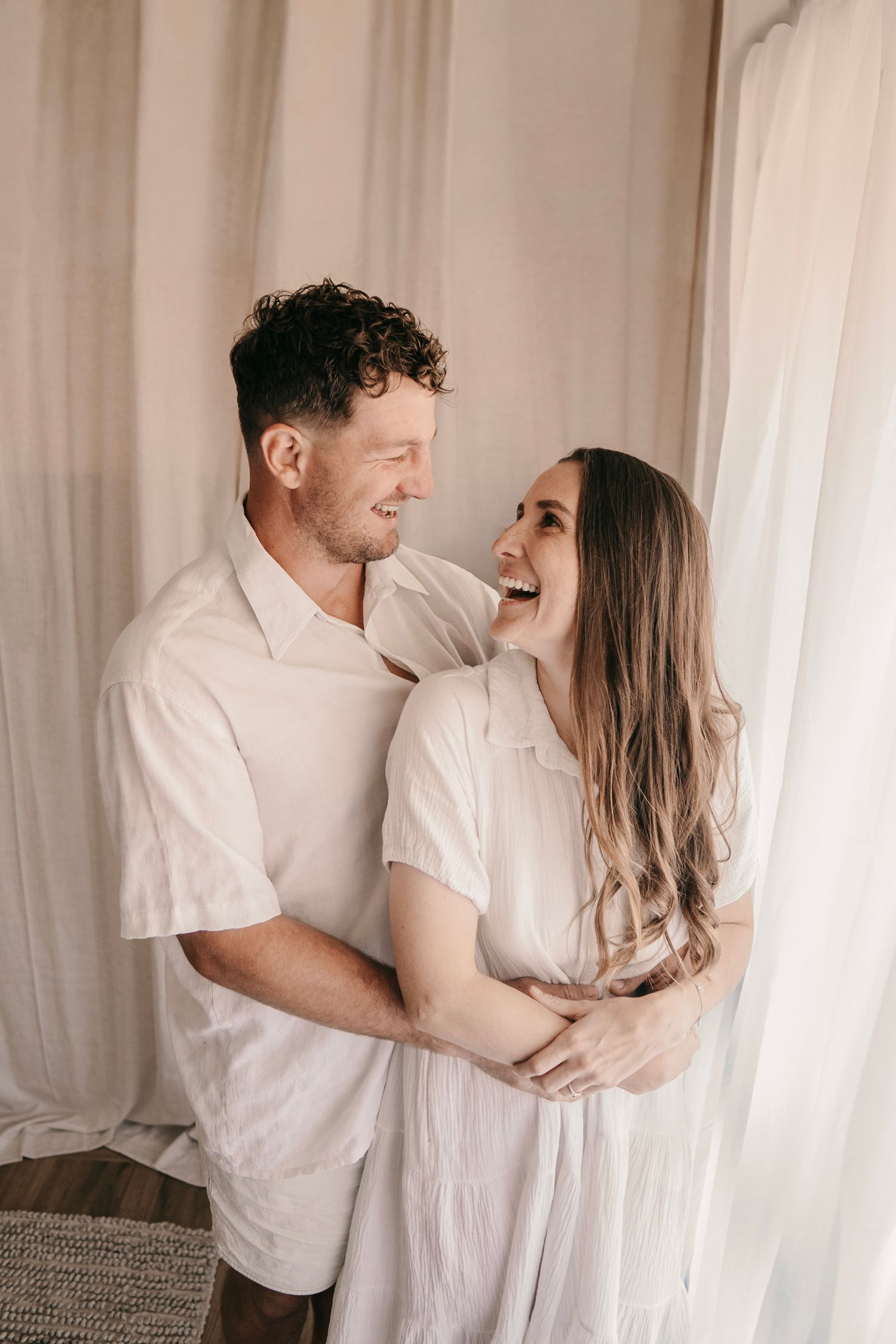 Happy couple in white outfits embracing and laughing together in a bright, airy room with sheer curtains.