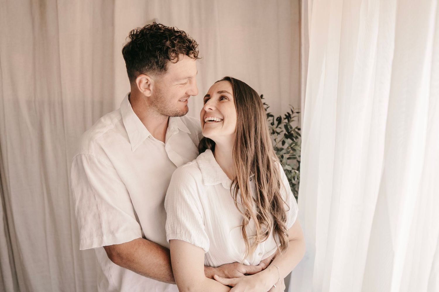 A happy couple embracing and smiling at each other in front of a soft white curtain backdrop.