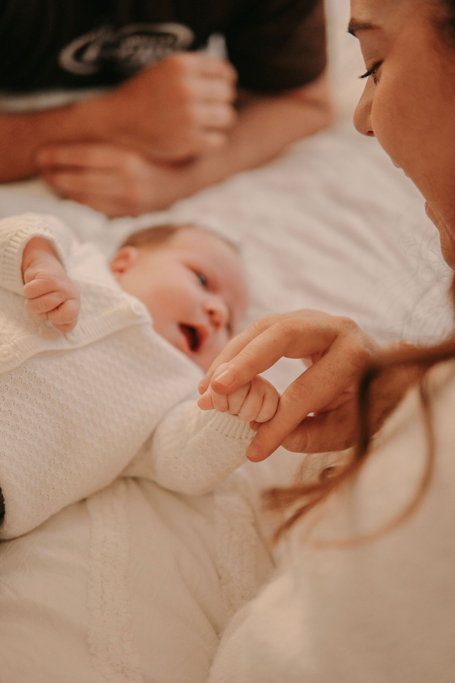 Mother gently holding her yawning newborn baby's hand while baby wears a white knit sweater.