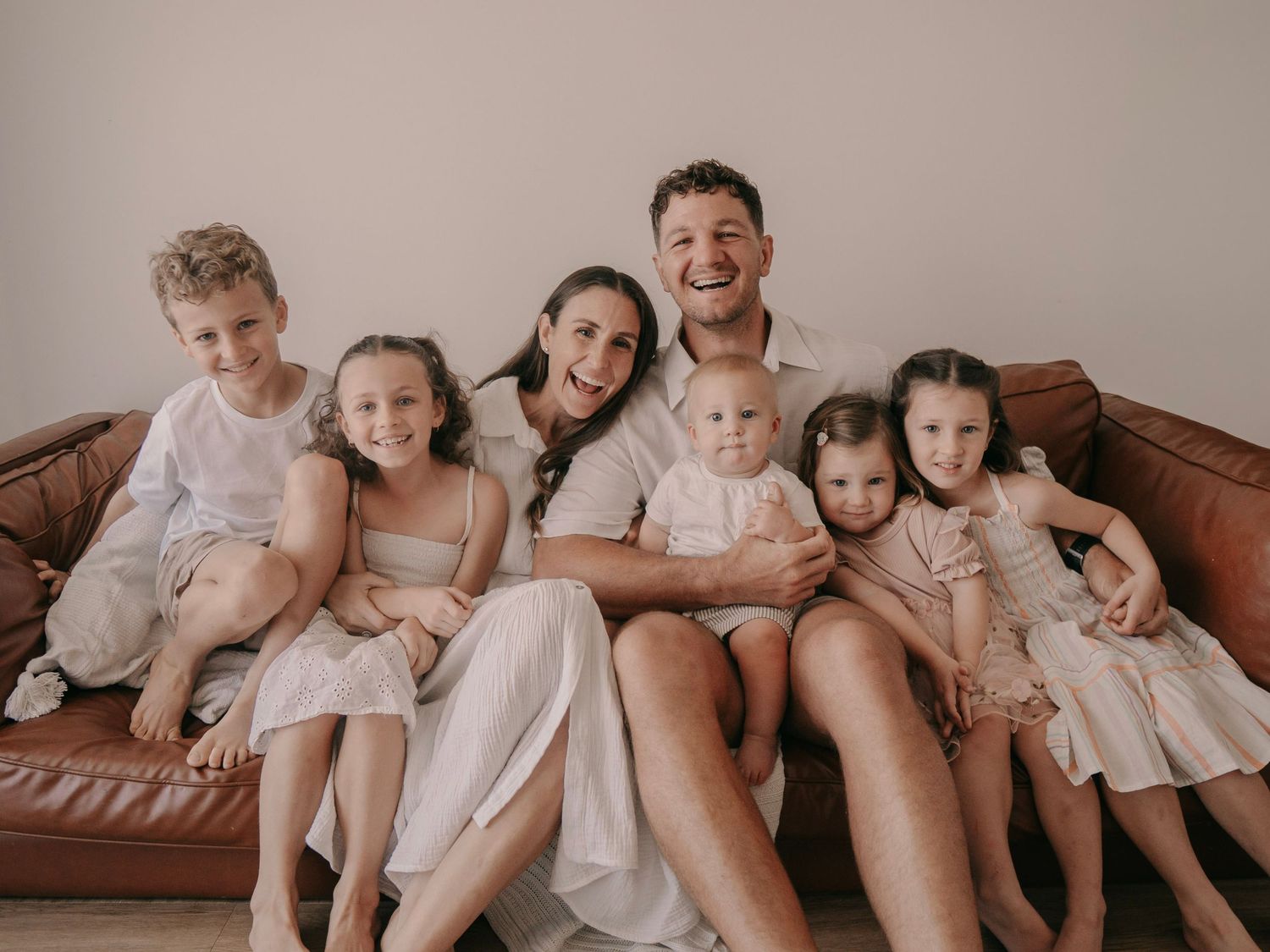 Happy family of seven posing together on a brown leather couch, wearing coordinating white and cream outfits.