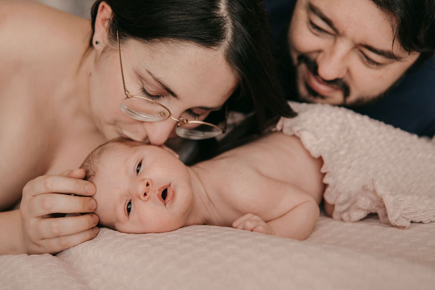 Parents lovingly kissing their newborn baby lying on a soft pink blanket in a tender family moment.