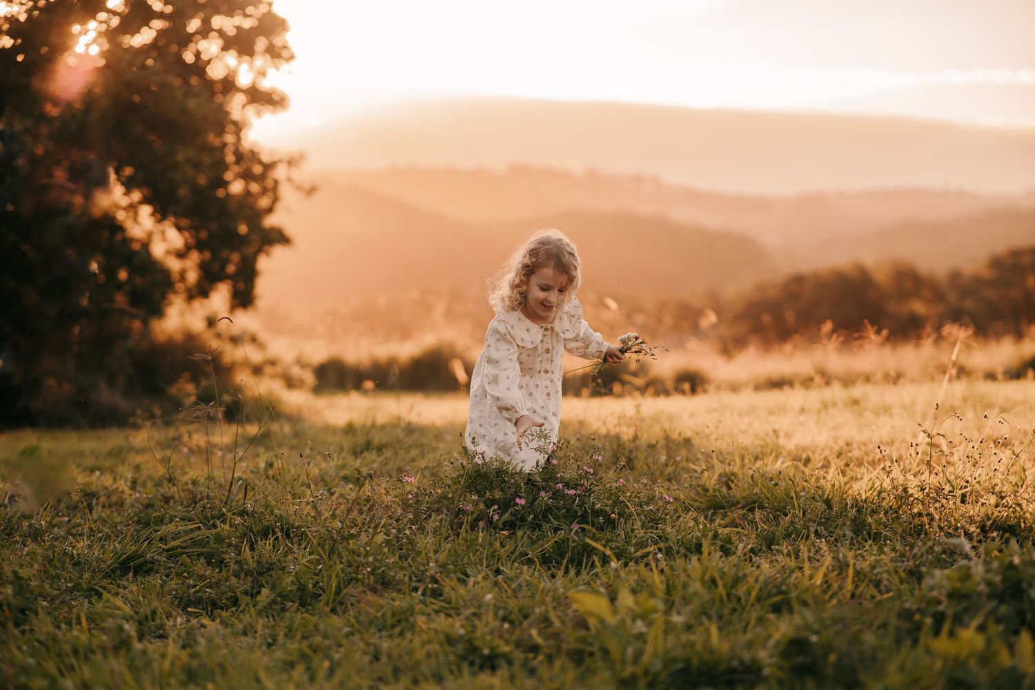 Adorable curly-haired toddler in white dress playing in a sunlit meadow with stunning mountain sunset backdrop.