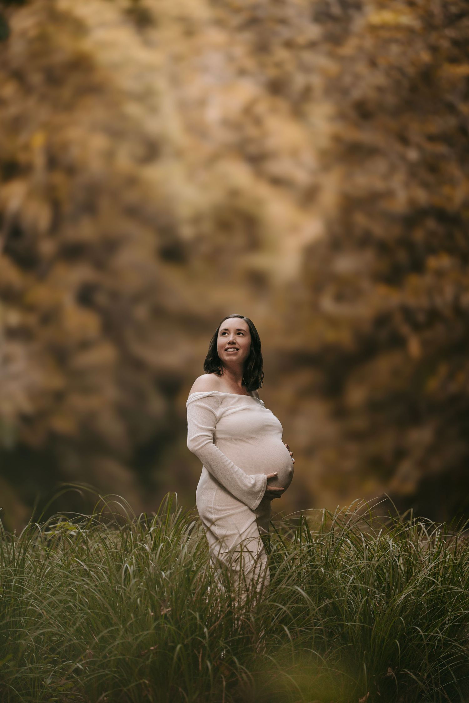 A pregnant woman in a white dress standing in tall grass with a rocky backdrop.