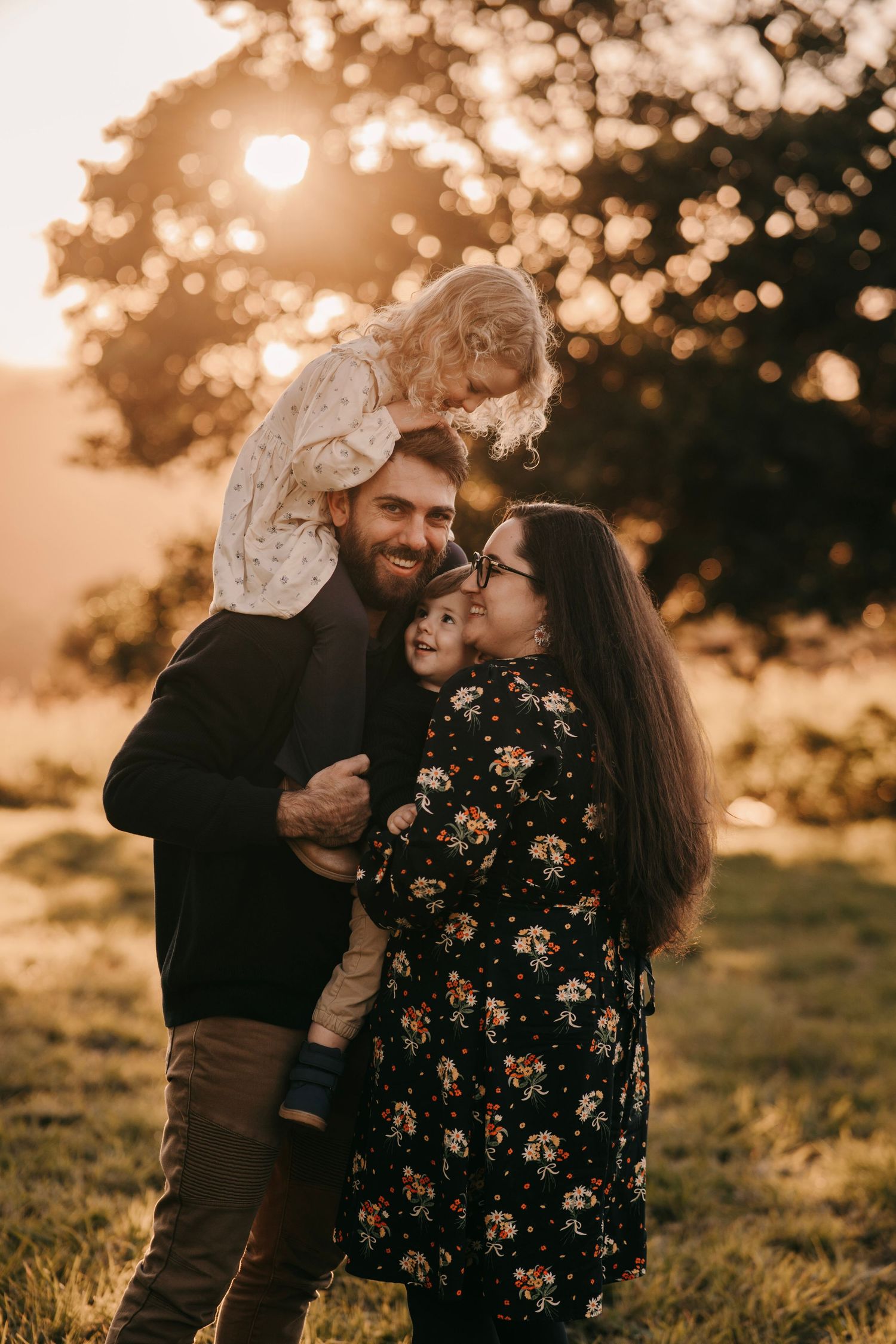 A happy family poses outdoors at golden hour, parents holding two young children in a sunlit park.
