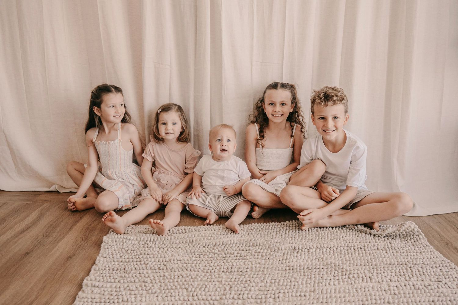 Five young children sitting together on a rug smiling in coordinated neutral outfits against a cream backdrop.