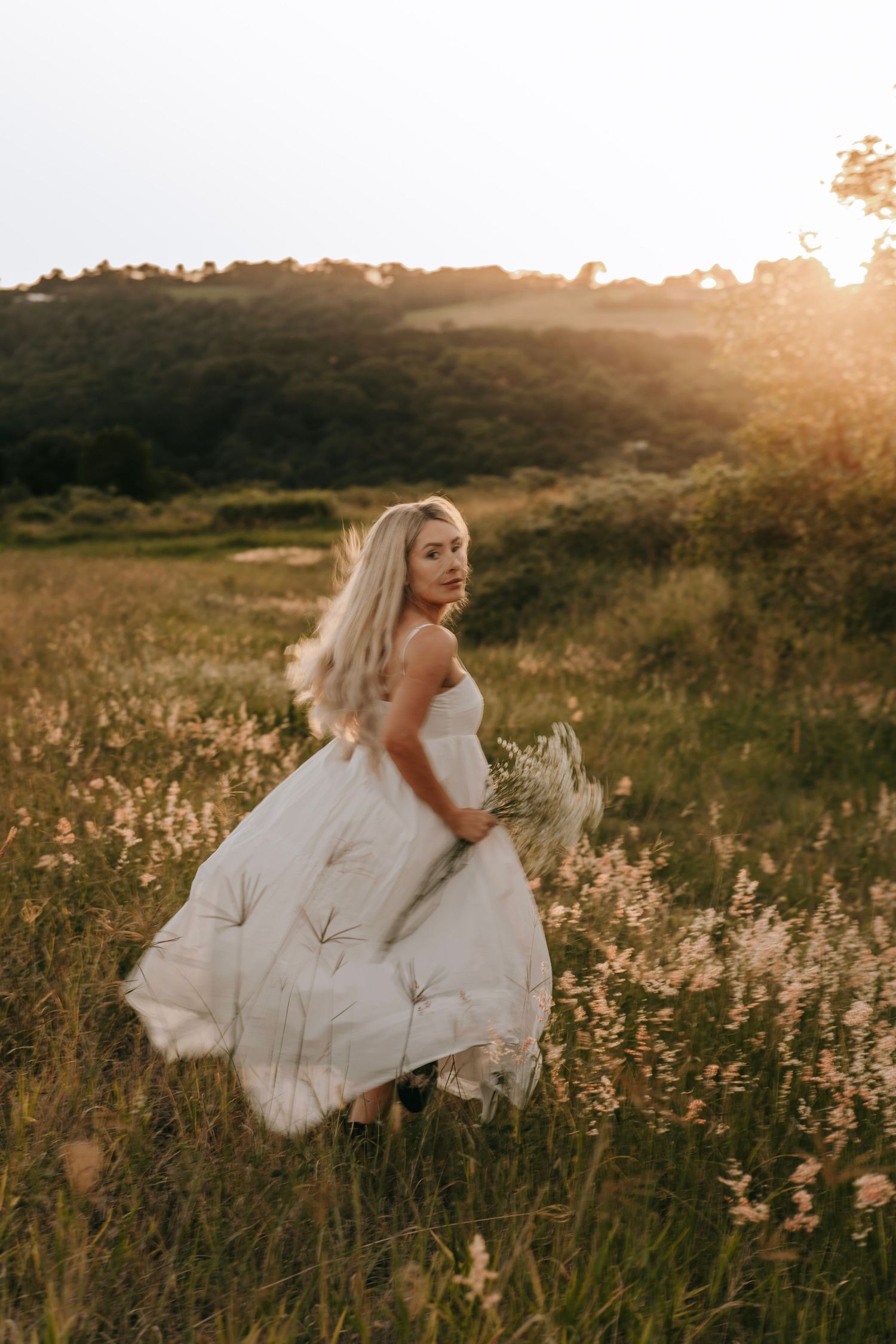 Blonde bride in white wedding dress running through a wildflower field at golden sunset.