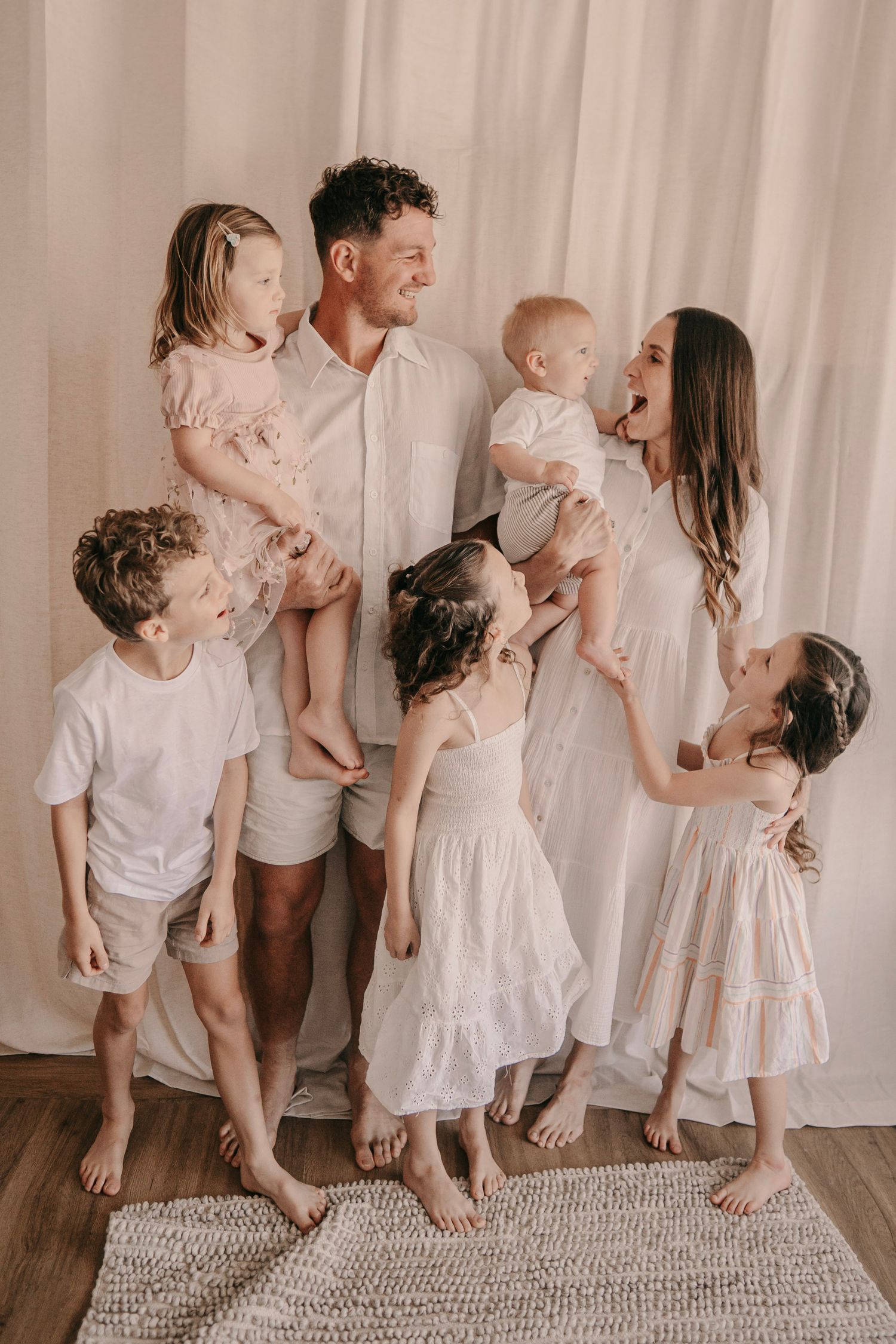 Happy family with five children posing together in white outfits against a light curtain backdrop in a bright studio.
