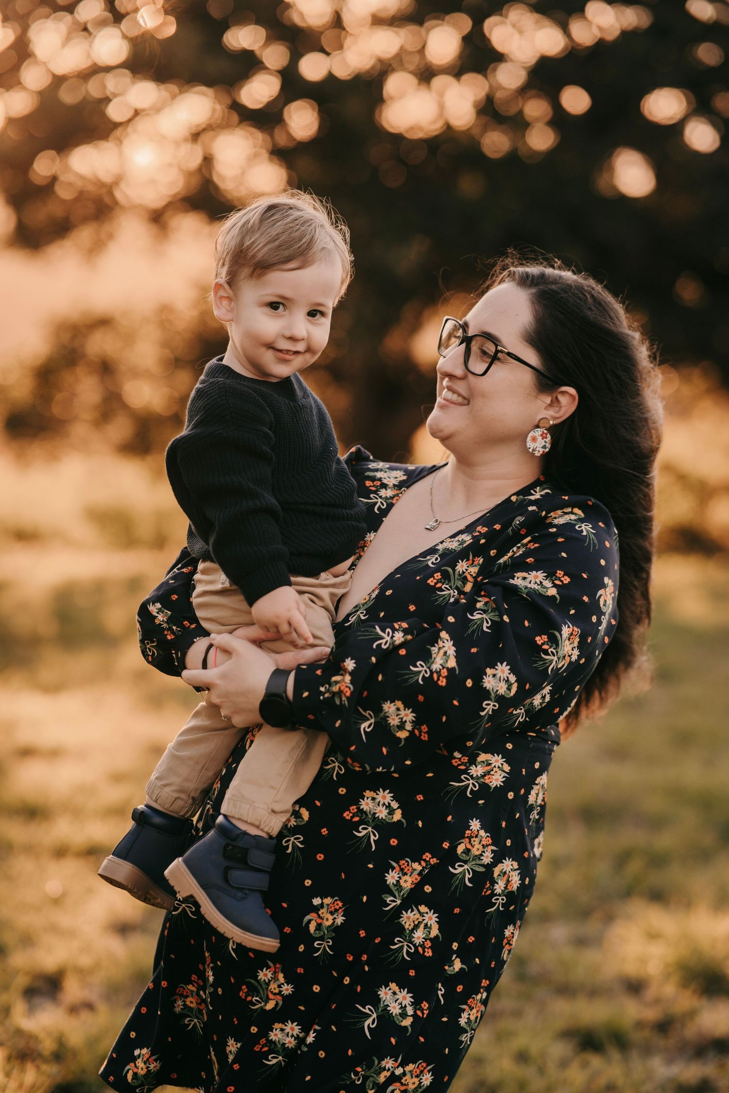 A smiling mother in a floral dress holds her young son during a warm sunset family portrait.