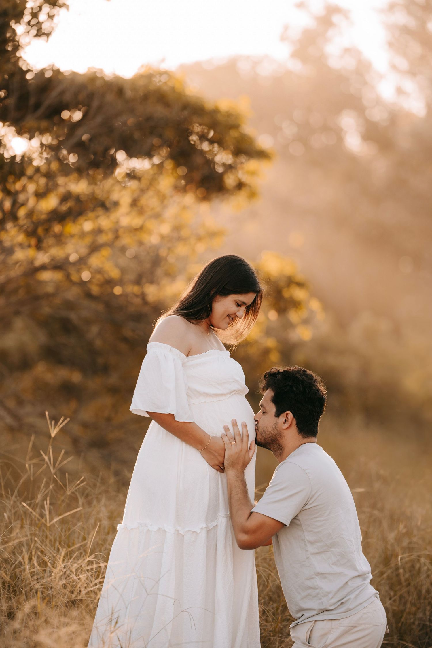 Man kneeling to kiss pregnant woman's belly in a sunlit outdoor field at golden hour.