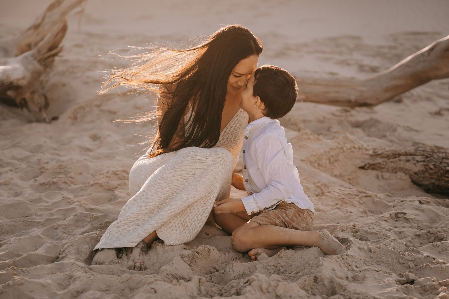 A mother and young son share a tender moment sitting together on sandy beach in warm golden light.