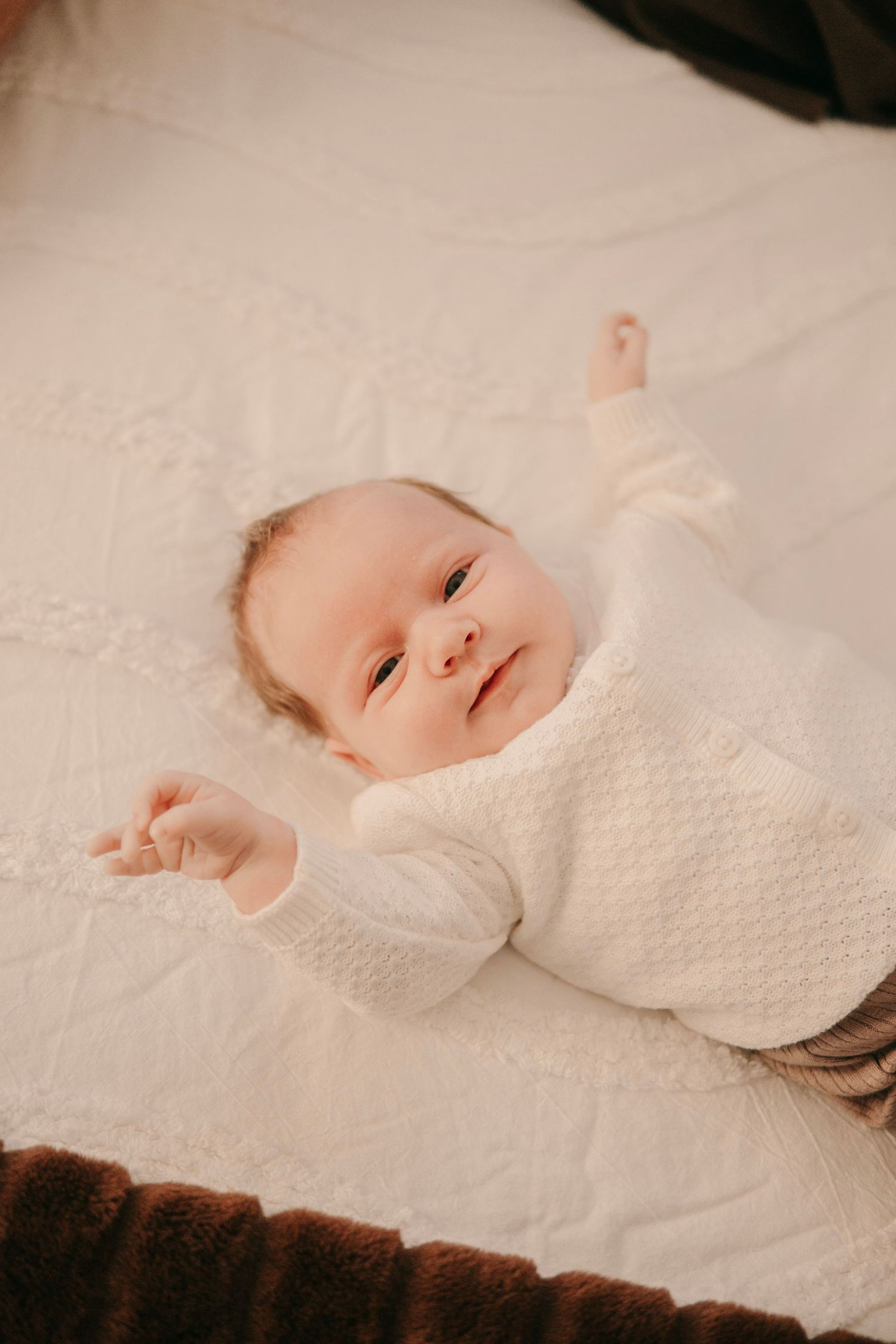 Adorable newborn baby in white knit outfit lying on a soft white blanket with arms stretched out.