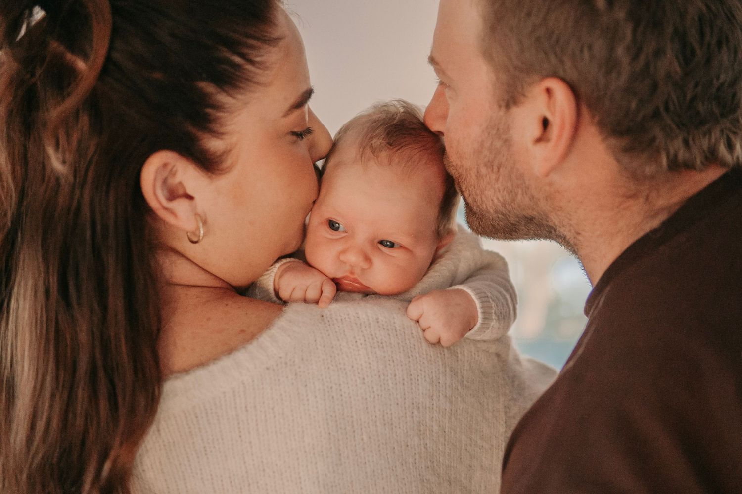 Parents kissing their newborn baby on cheeks in a heartwarming family portrait.