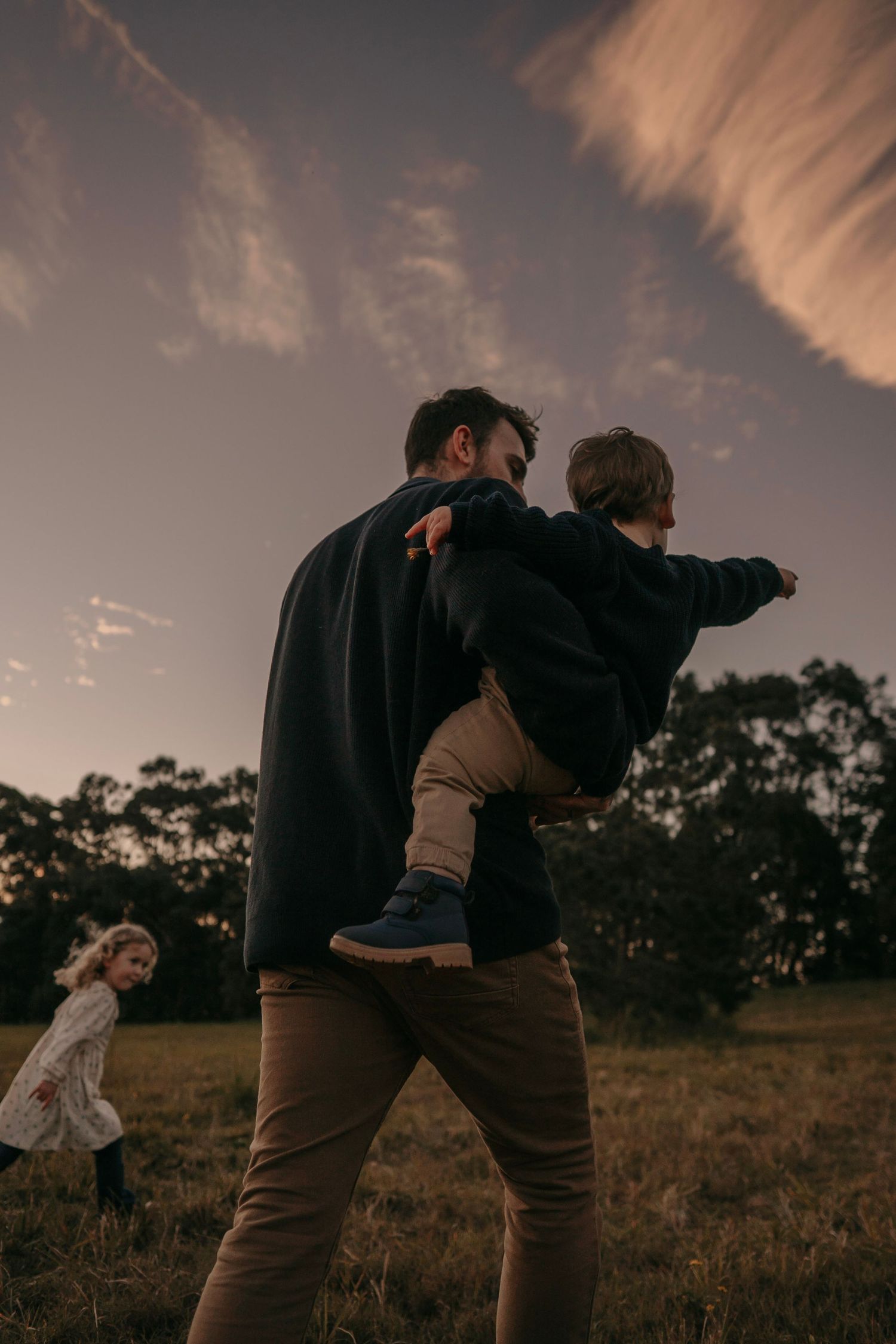 A father playfully carries his young son like an airplane in an open field at dusk.