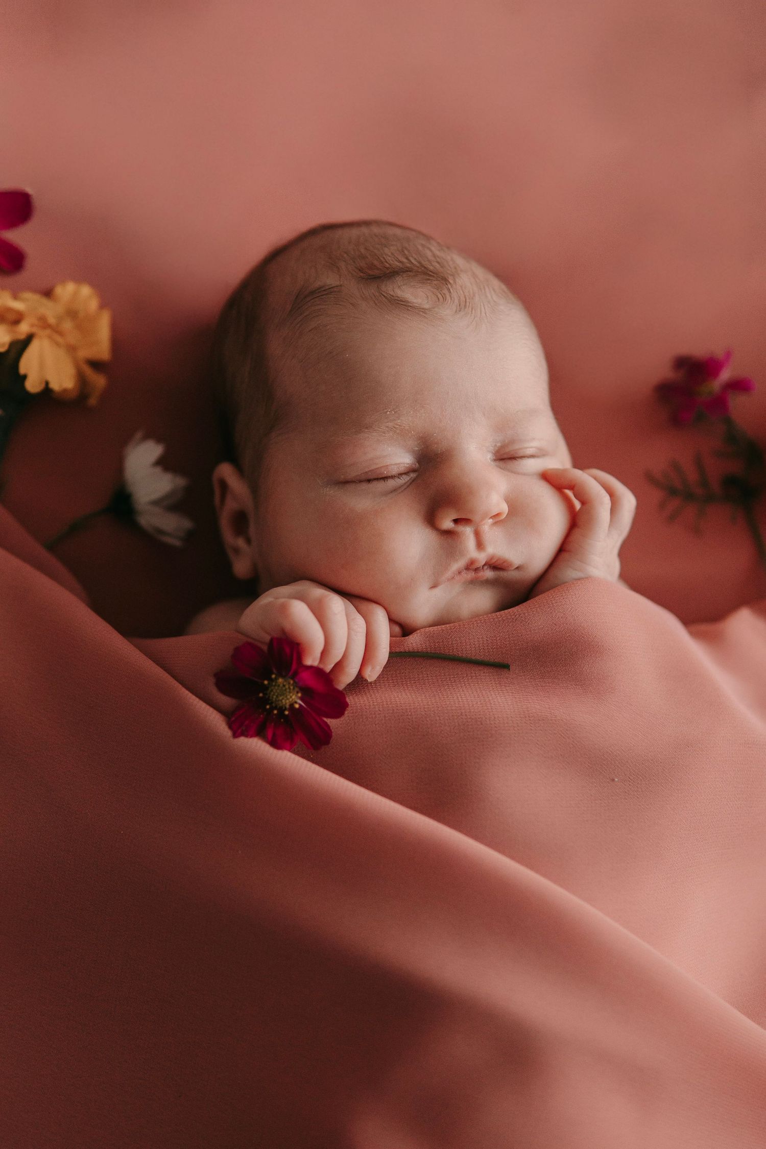 Peaceful newborn baby sleeping on pink fabric surrounded by colorful flowers.