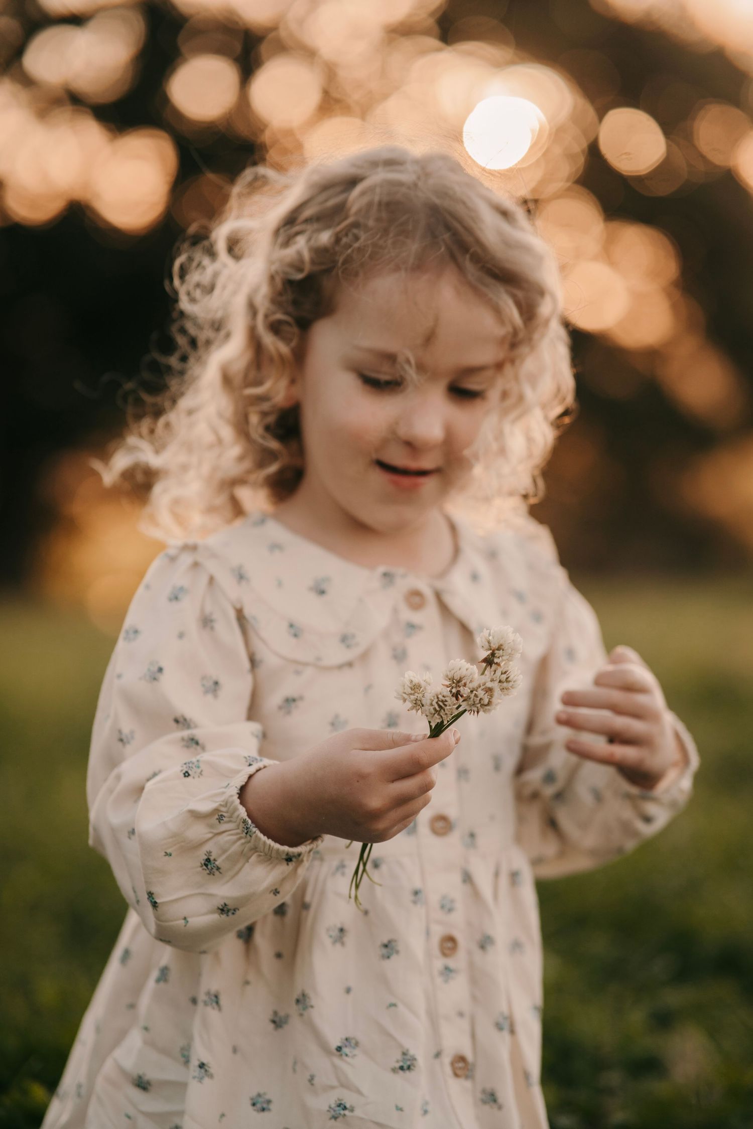 A curly-haired little girl in a floral dress holds wildflowers in a sunlit outdoor setting.