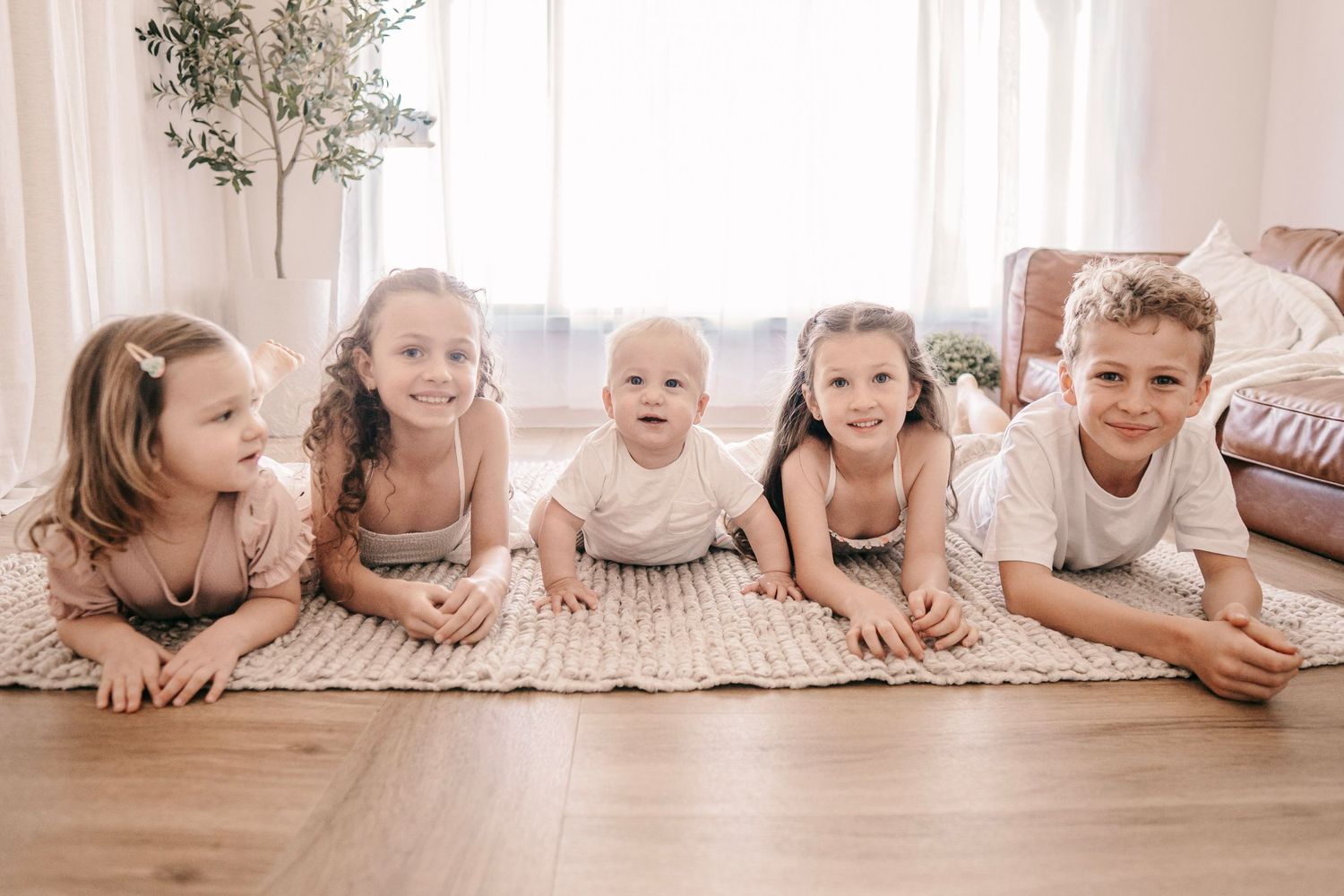 Five young children lying on a cozy rug smiling at the camera in a bright, airy living room.