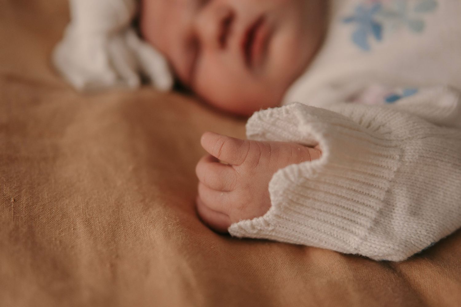 Sleeping newborn baby in white knit outfit with tiny hand resting on blanket.