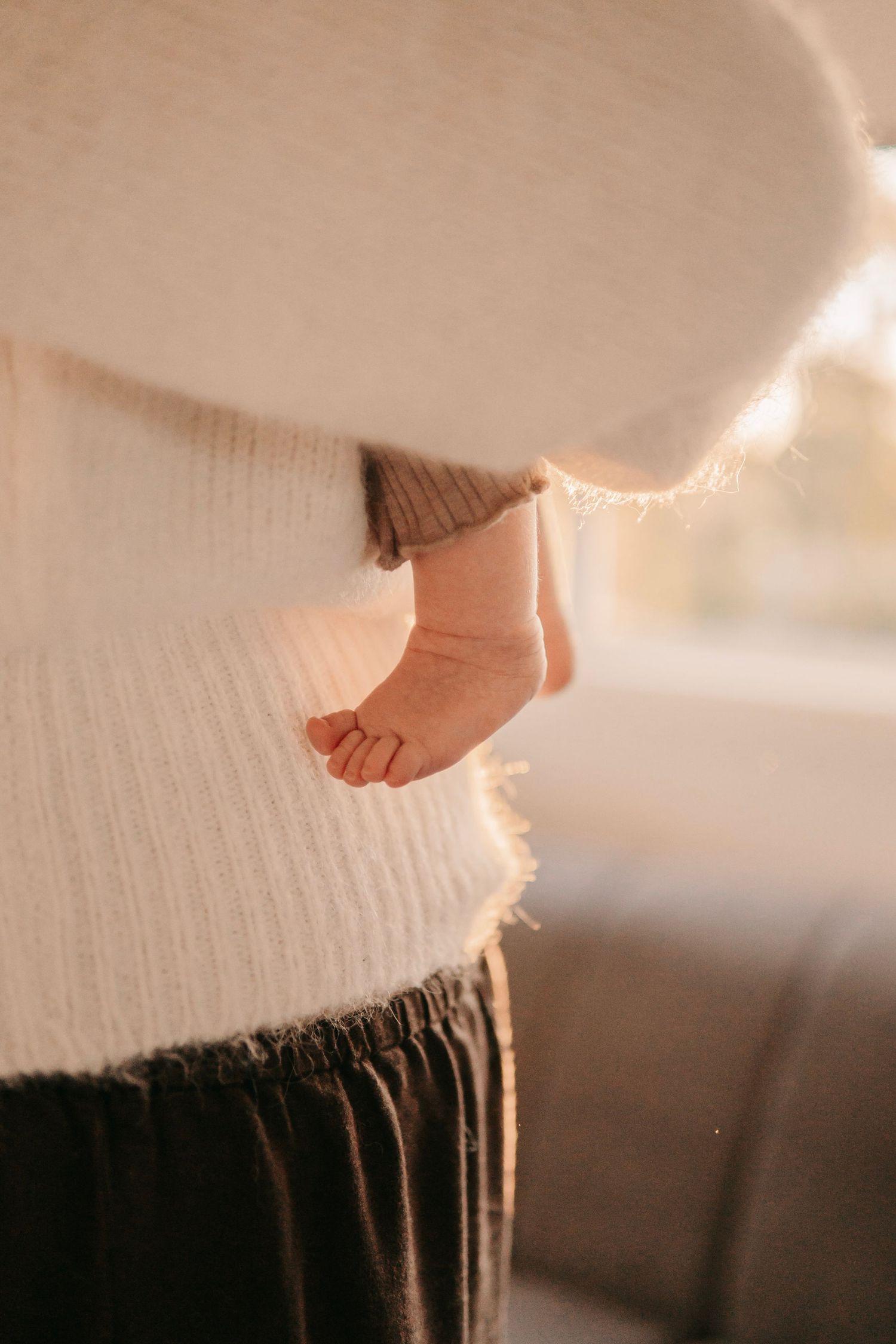 Newborn babys tiny feet peeking out from mothers cozy white knit sweater in warm light.