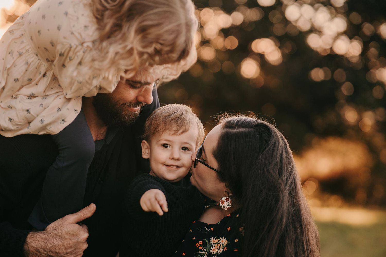 Family of three sharing a tender moment outdoors, parents kissing toddler boy in warm golden light.