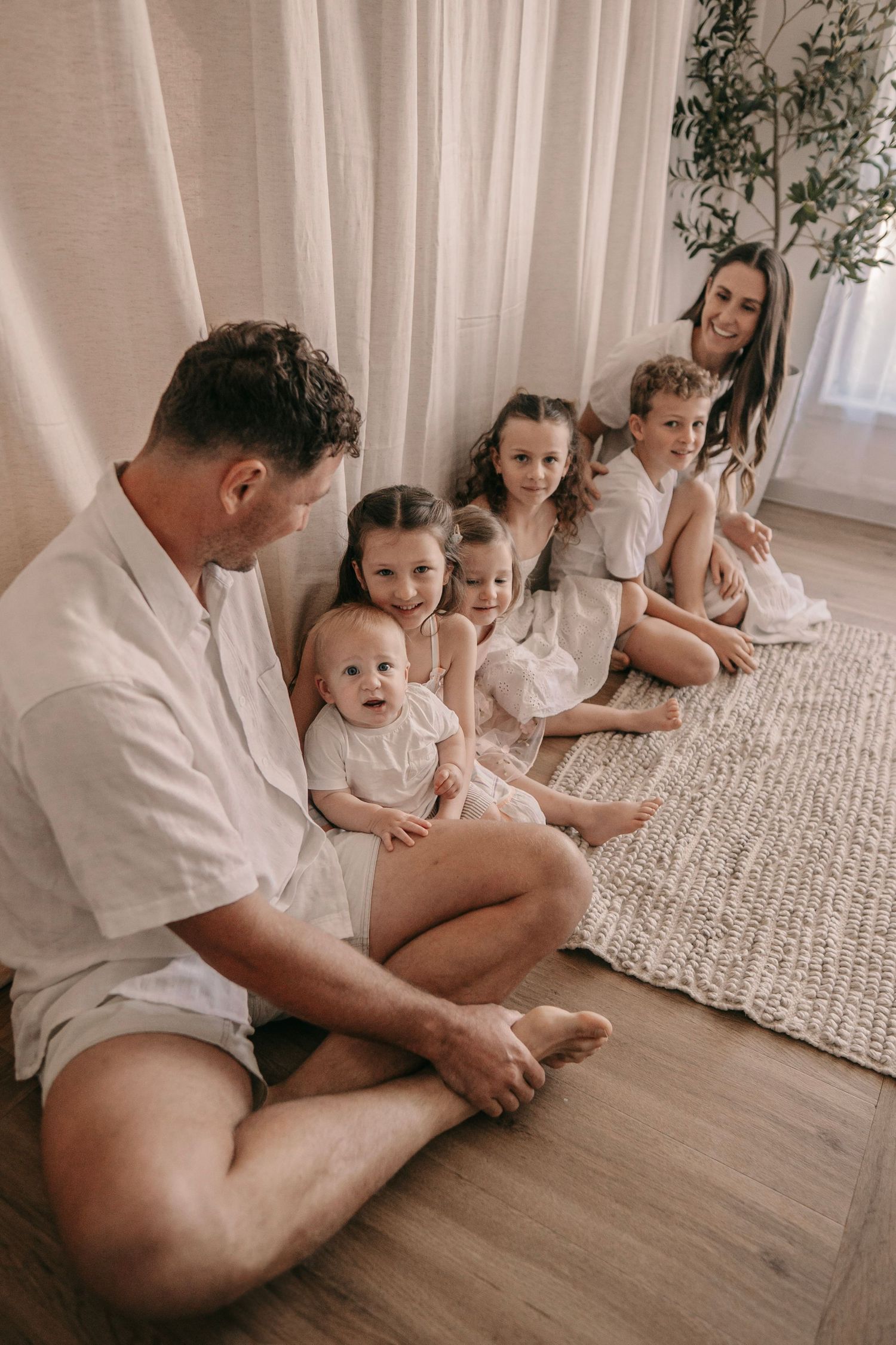 A family of seven in white clothing sitting together on a wooden floor and rug in a bright, cozy room.