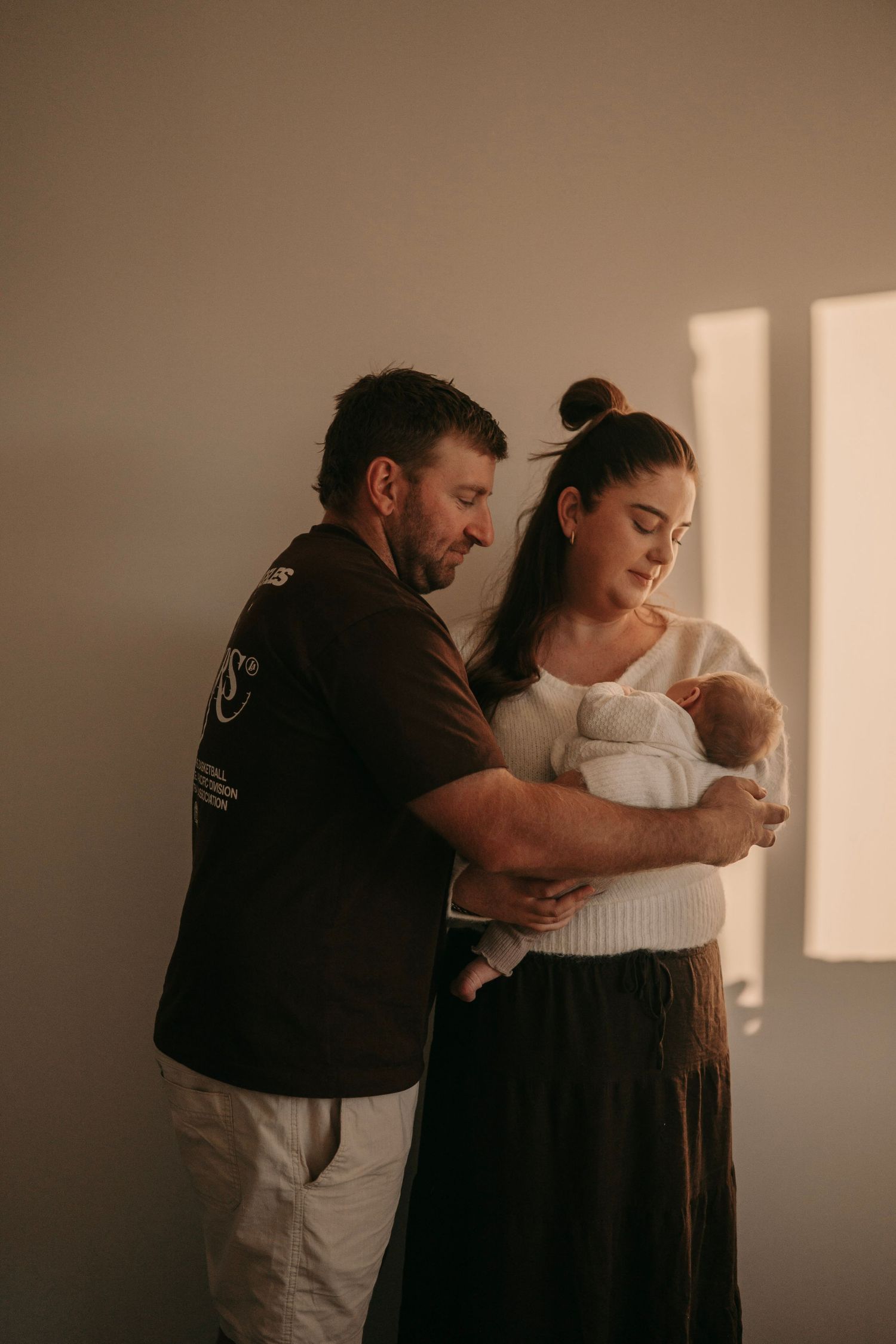 A couple standing by a sunlit window tenderly holding their newborn baby wrapped in white.