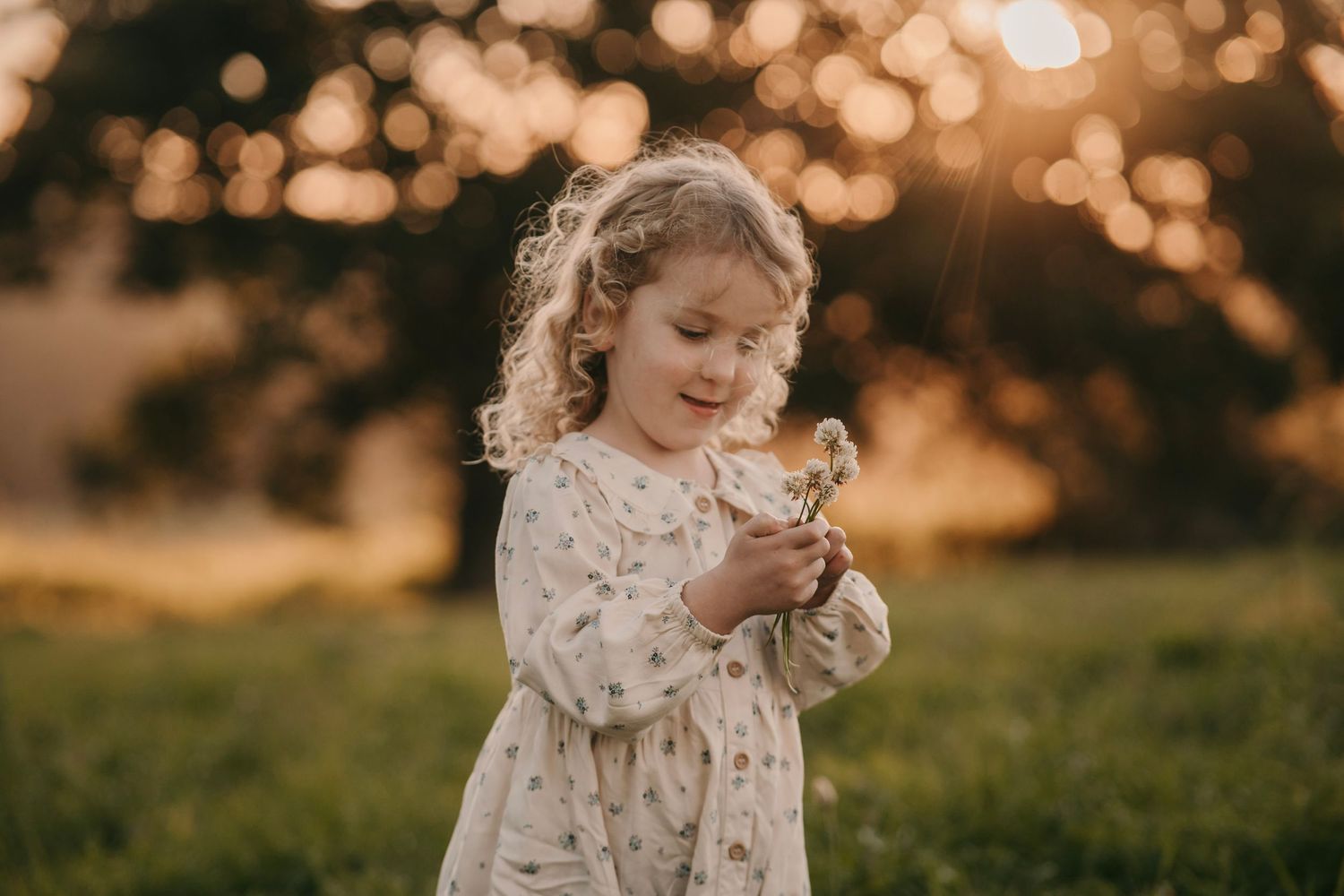 Adorable curly-haired toddler girl in floral dress holding flowers at sunset.