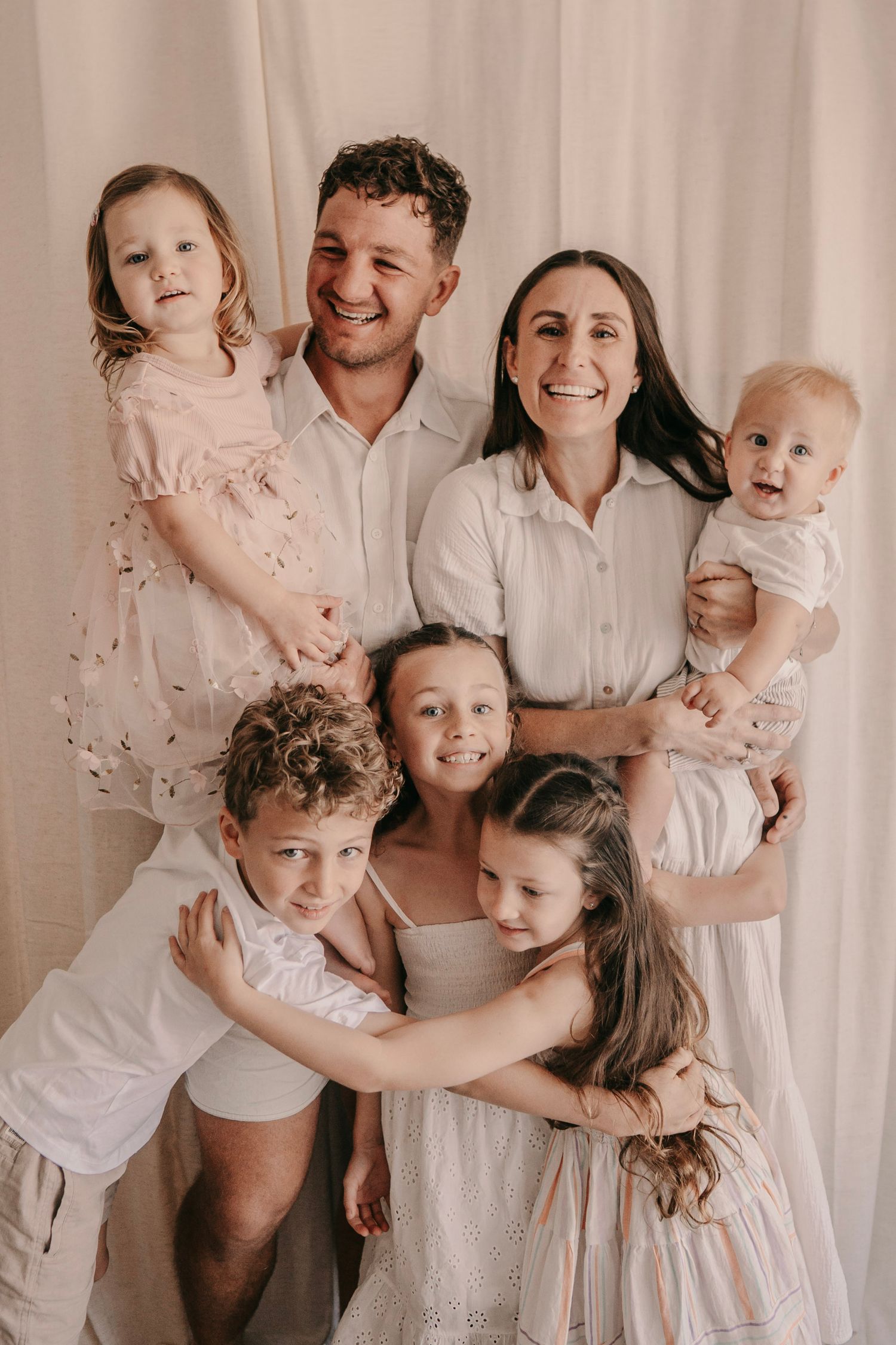 Happy family portrait with parents and five young children embracing, dressed in white and blush tones against a neutral backdrop.