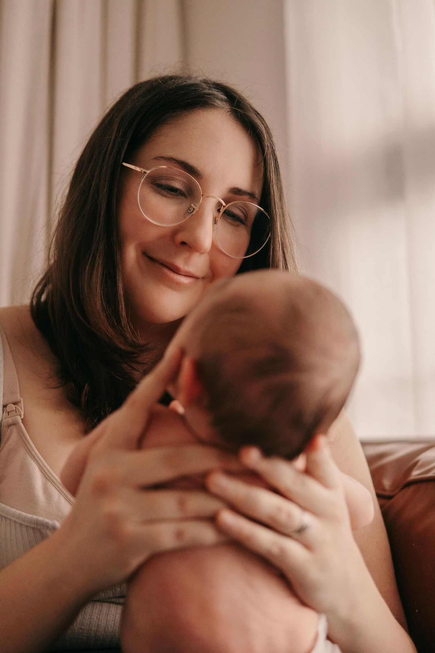 Smiling mother with glasses tenderly holding her newborn baby in soft natural light.