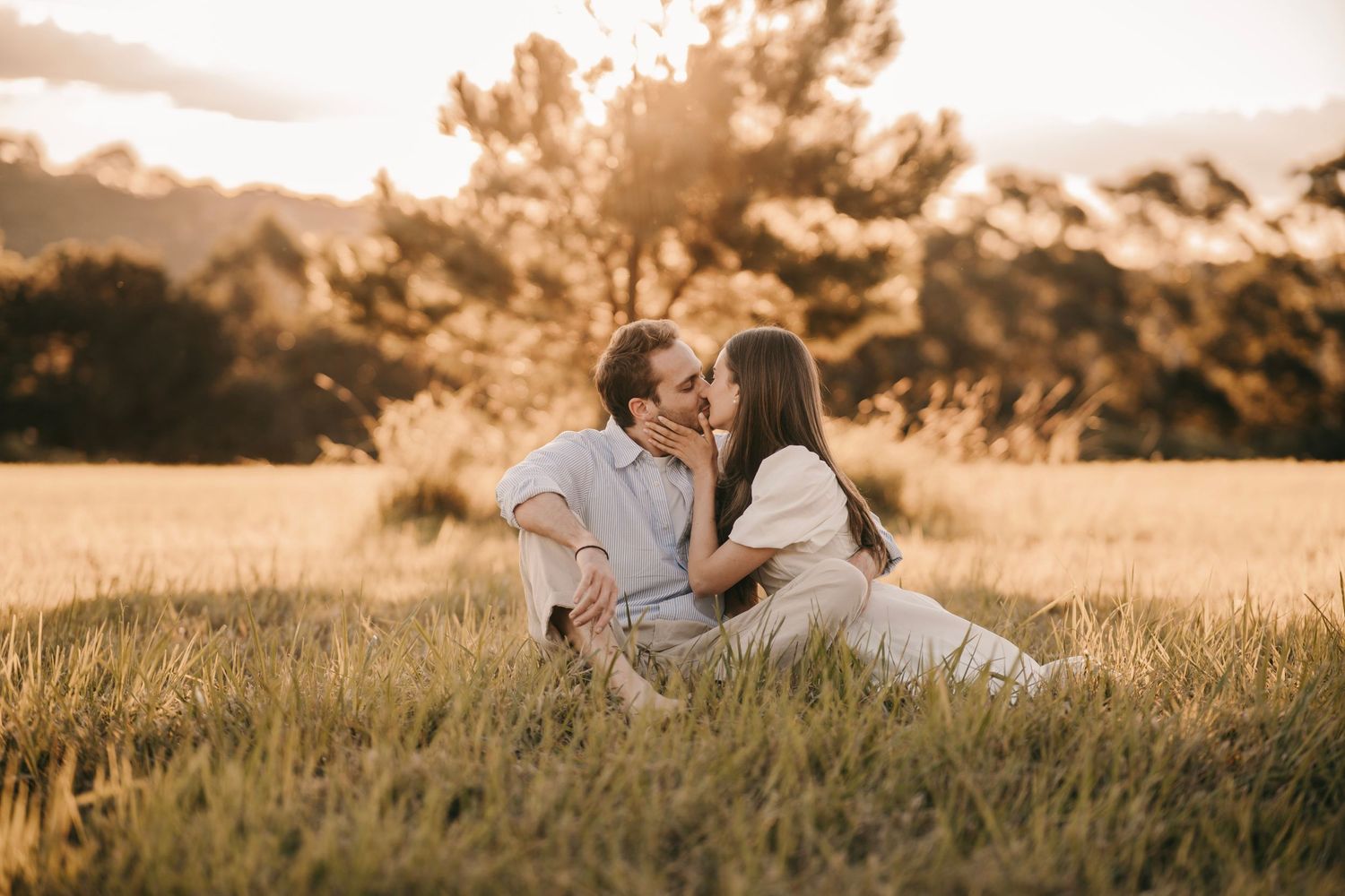 A romantic couple sharing a kiss while sitting in a lush golden grass field at sunset.