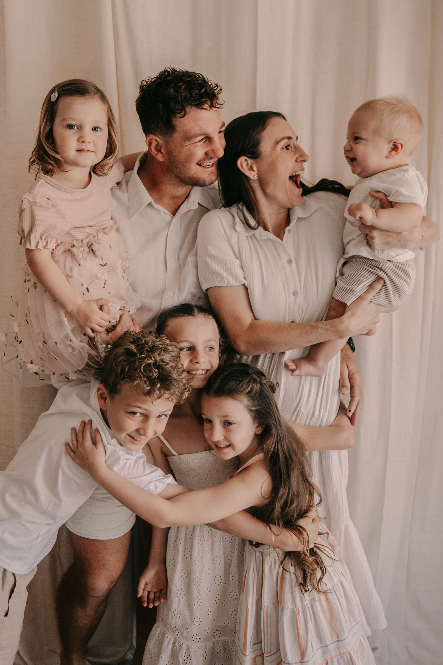 Happy family portrait with parents and five young children embracing and laughing against a soft white curtain backdrop.