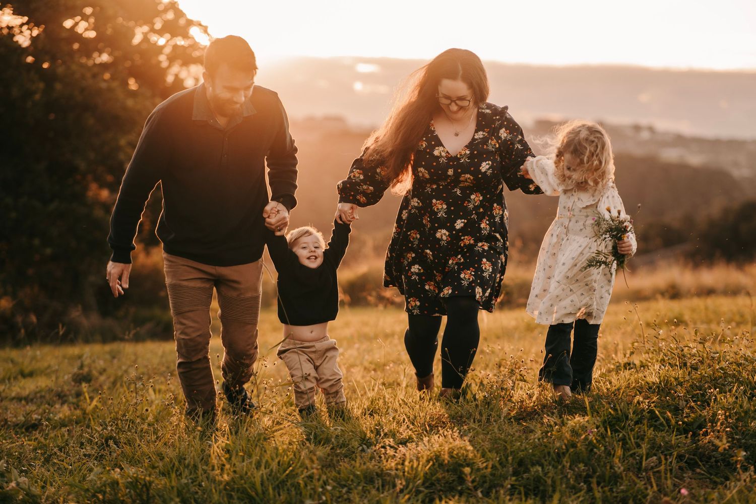 Family of four walking hand-in-hand through golden field at sunset, parents swinging young children between them.