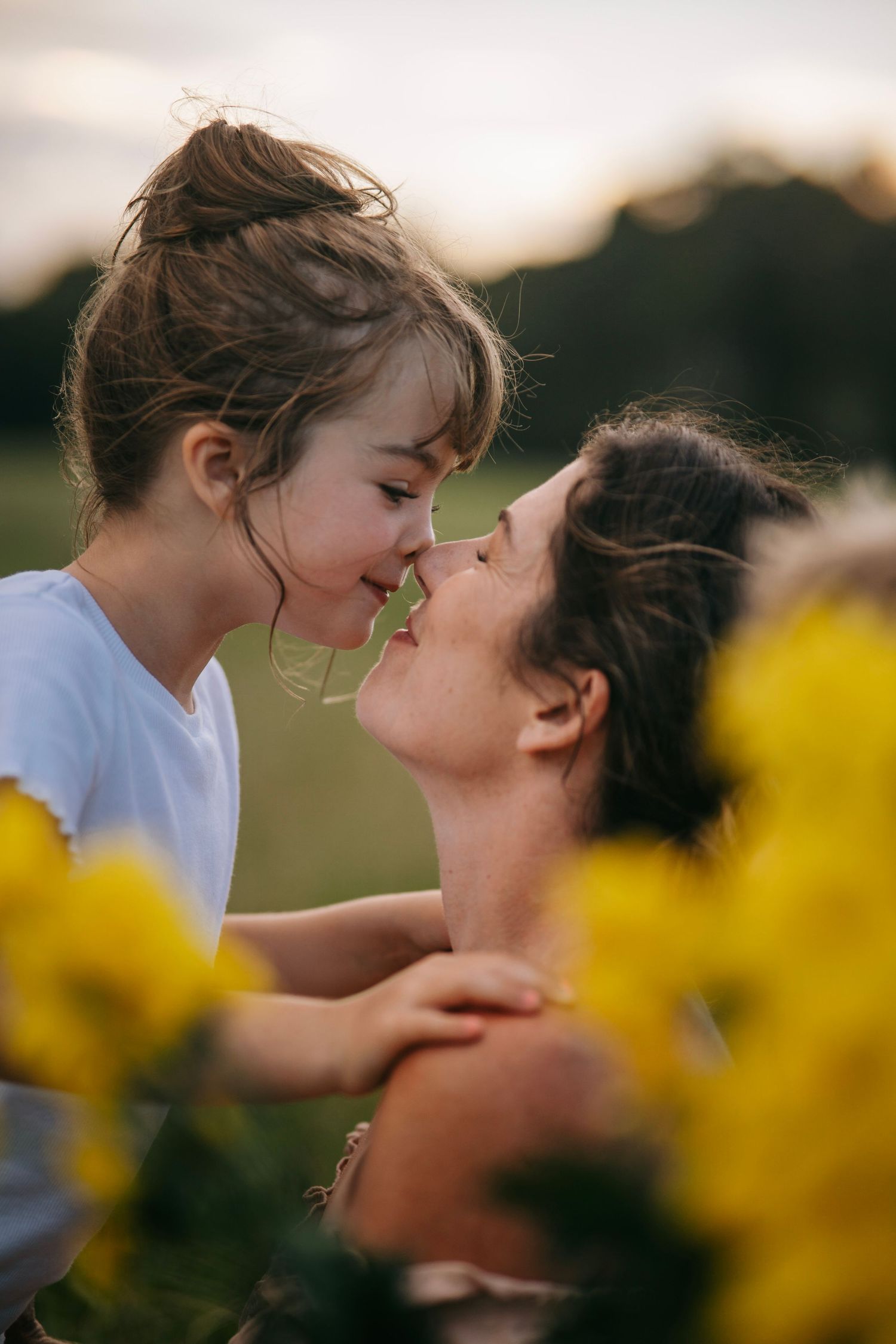 A mother and young daughter share a tender nose-to-nose moment surrounded by yellow flowers in a sunlit field.