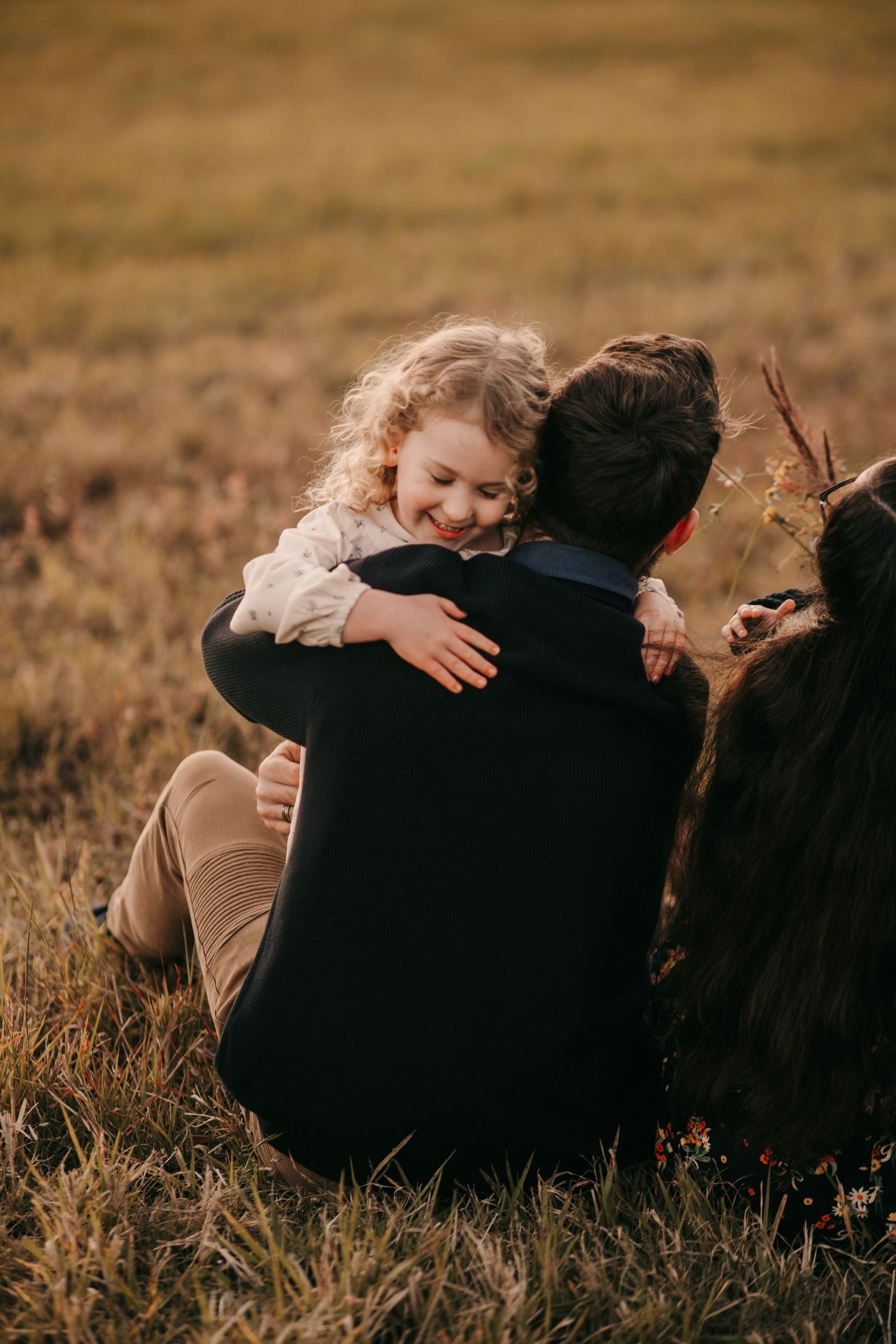 Joyful curly-haired girl hugging her father tightly while sitting in an autumn field.