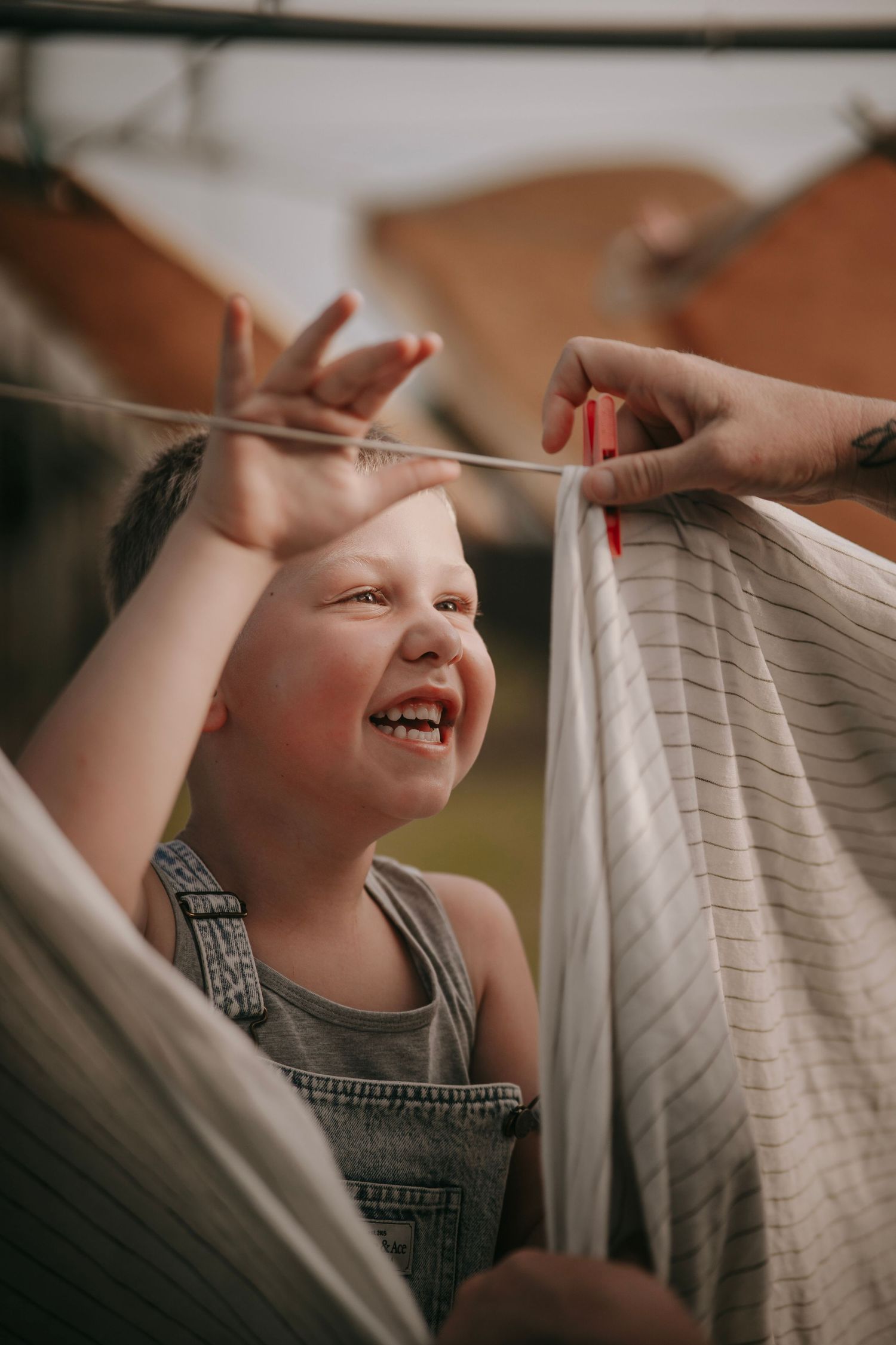 Happy laughing toddler in denim overalls helping hang laundry on clothesline outdoors.