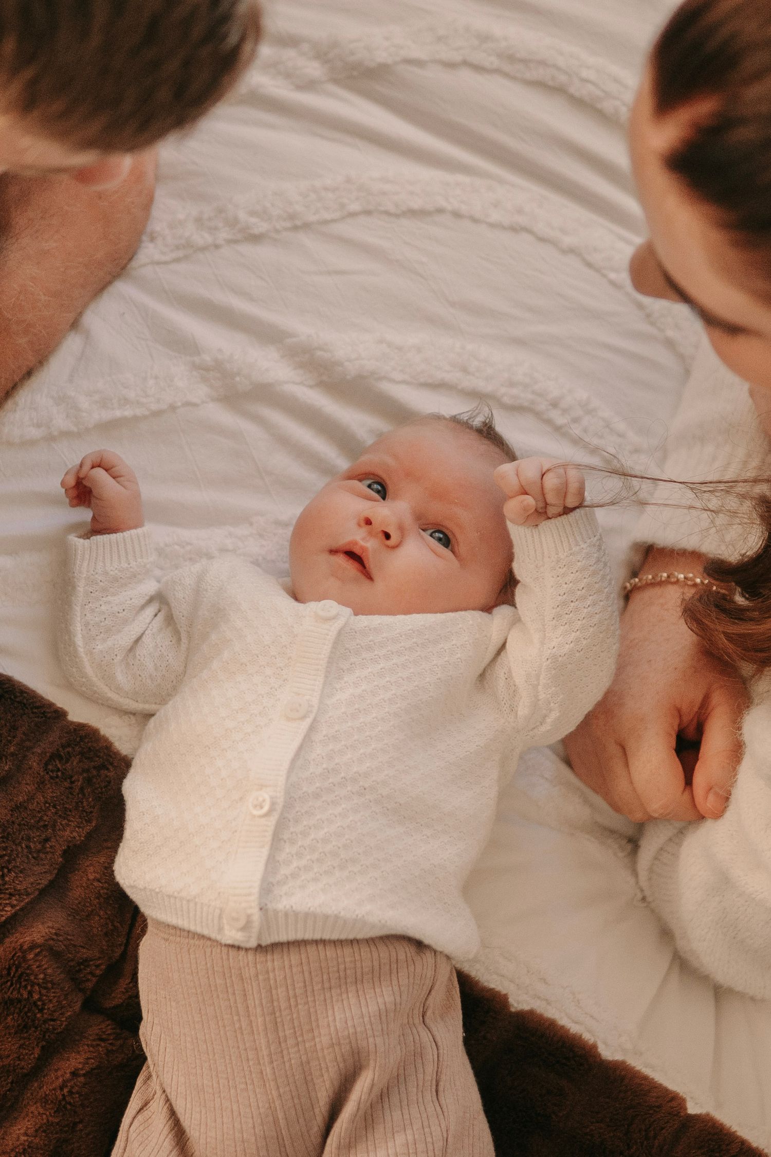 Parents lovingly gazing at their newborn baby dressed in a white knit cardigan on a white blanket.