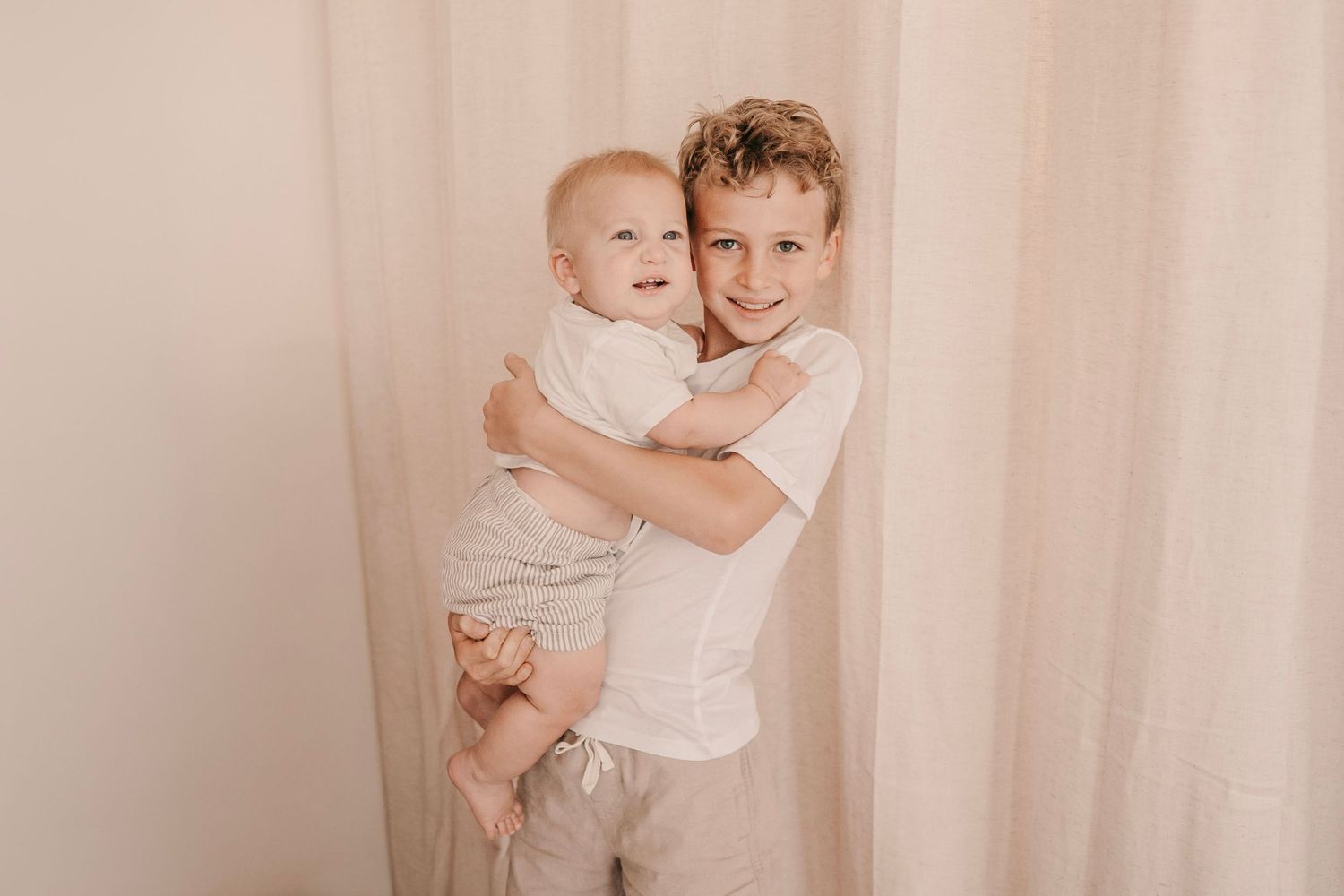 A young boy in a white shirt lovingly holds a smiling baby in front of soft beige curtains.